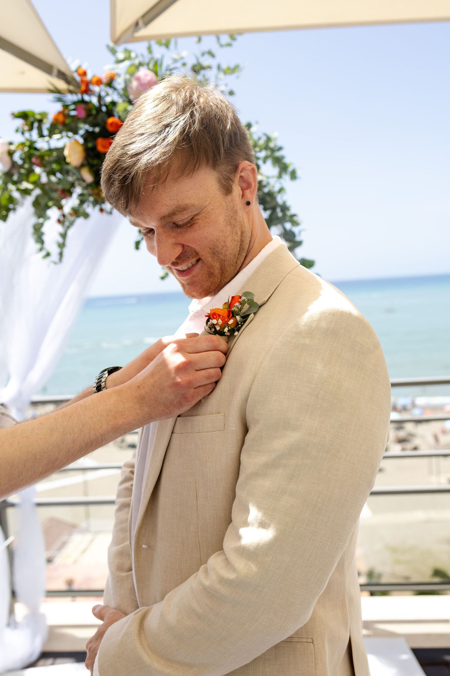 A smiling groom in a beige suit gets his boutonniere pinned near a seaside wedding arch.