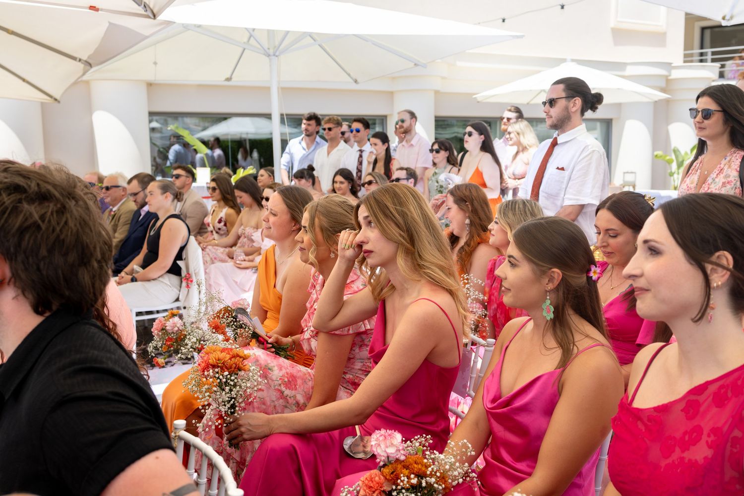 Bridesmaids in pink and orange dresses sit together smiling during an outdoor wedding ceremony.
