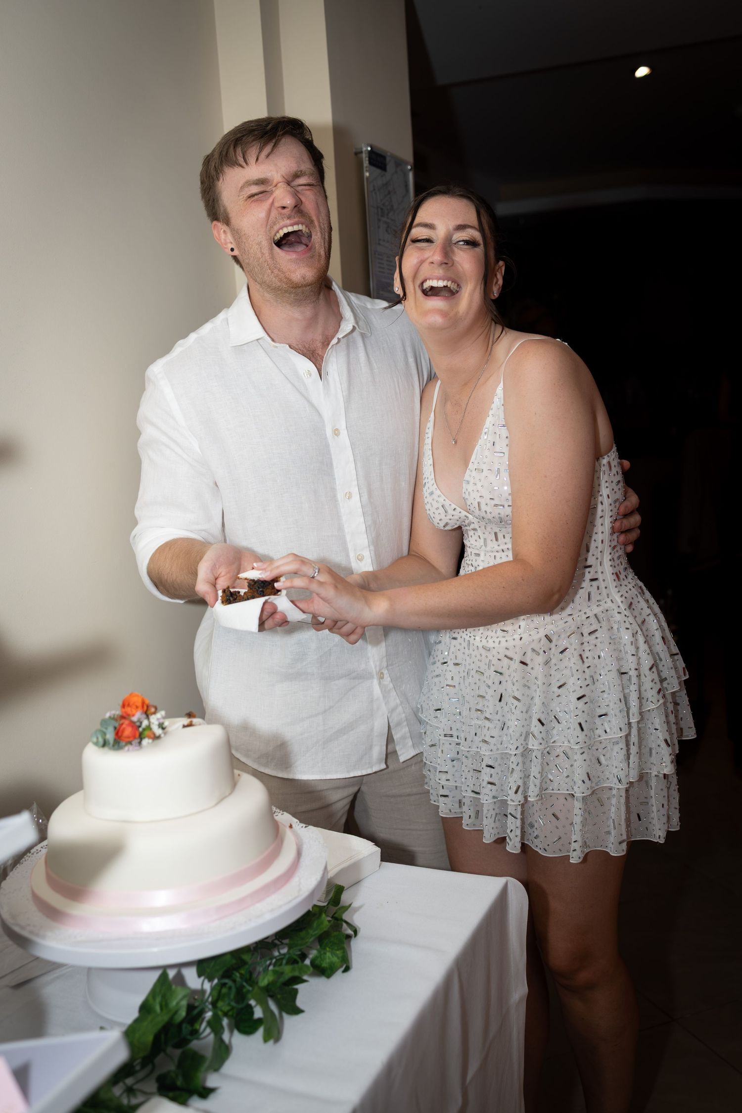 Laughing newlywed couple cutting their two-tier white wedding cake at their reception celebration.