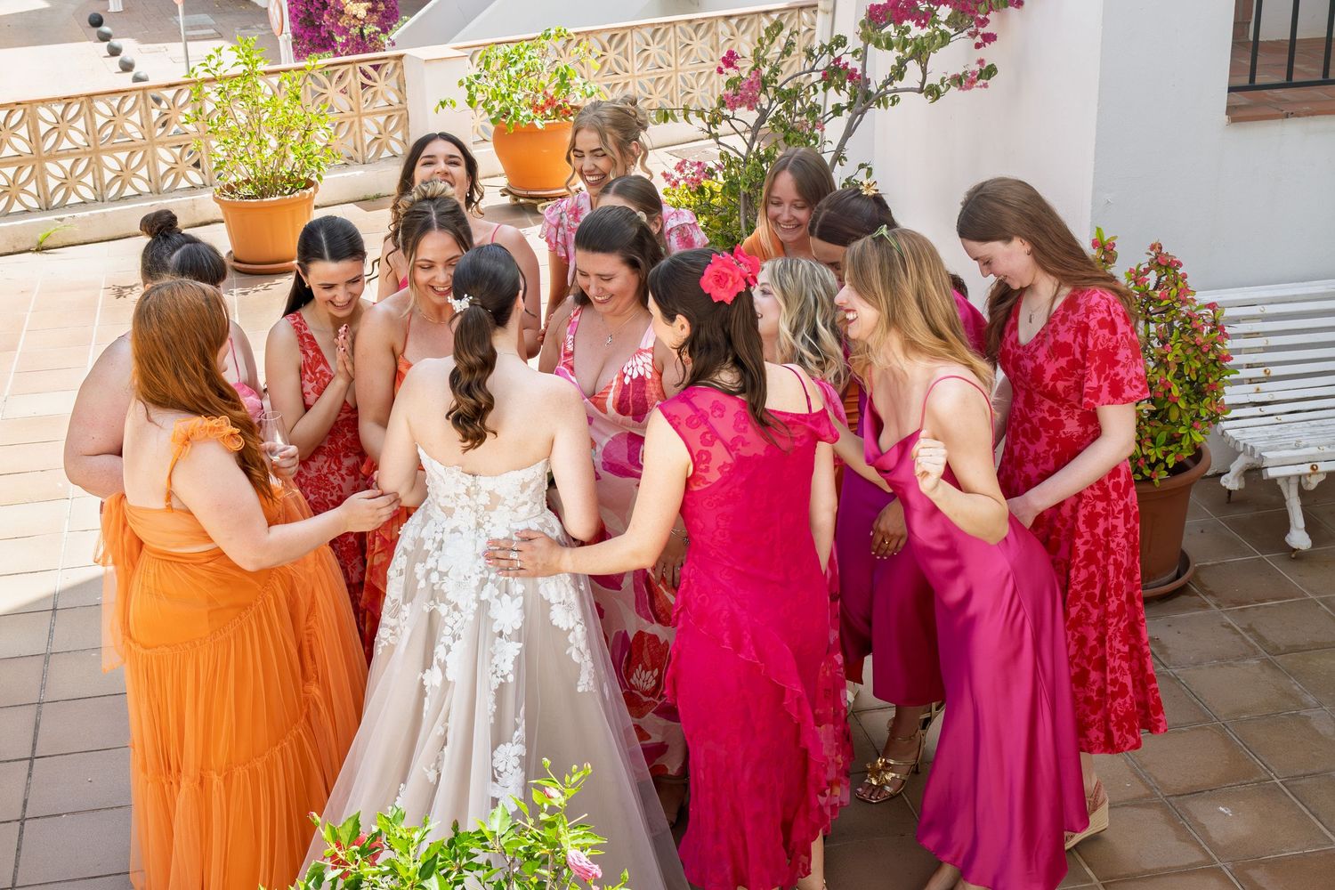 Bride in white lace gown surrounded by bridesmaids in vibrant pink and orange dresses on a sunny terrace.