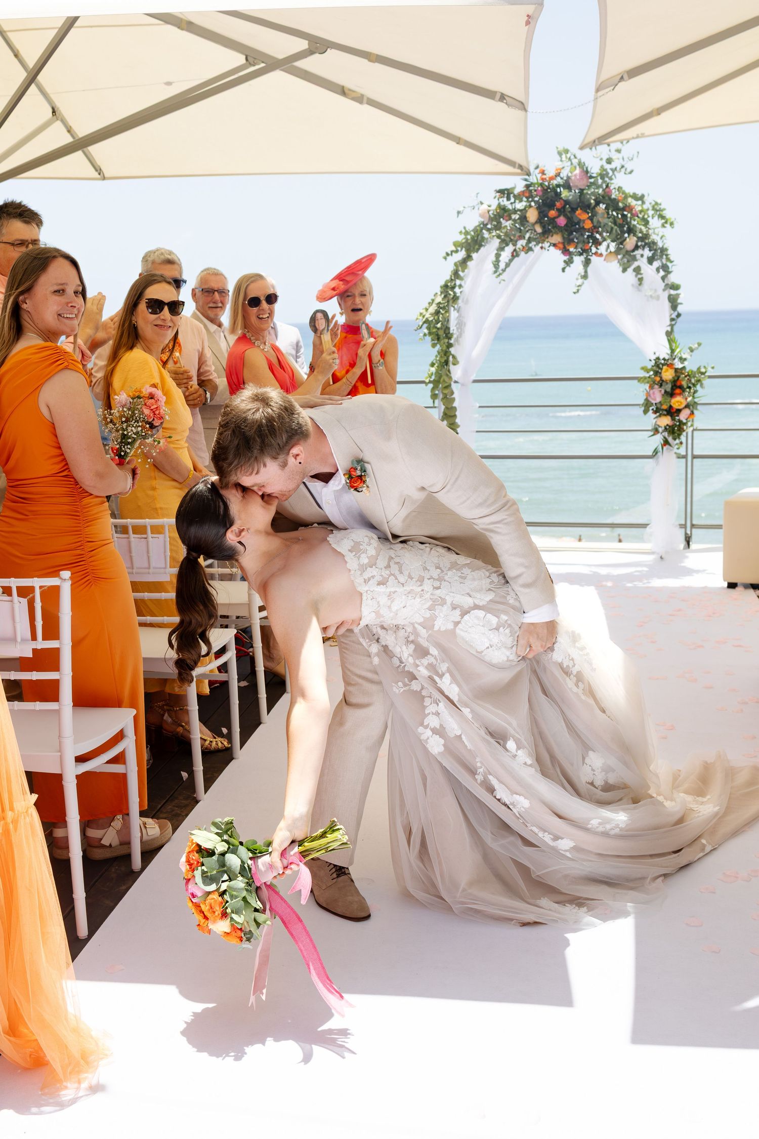 Groom dips bride for a romantic kiss at outdoor beach wedding ceremony with orange-dressed bridesmaids watching.