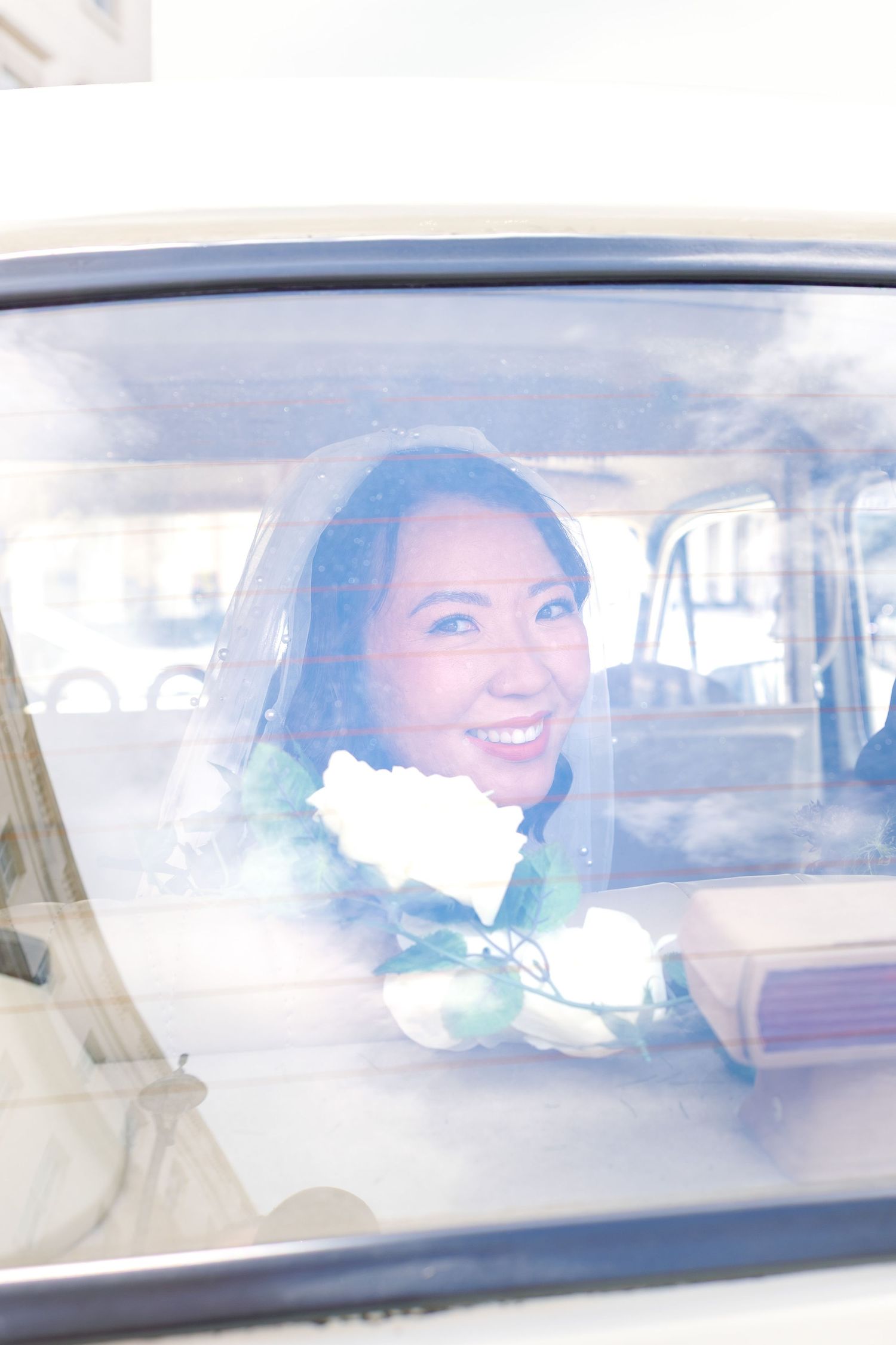 Smiling bride in veil holding bouquet seen through a vintage car window.