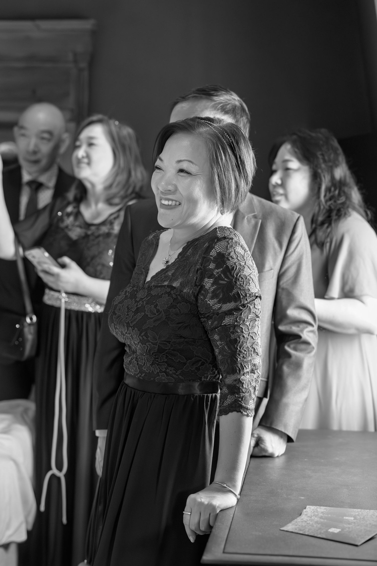 Smiling Asian woman in black lace dress at wedding ceremony surrounded by guests.