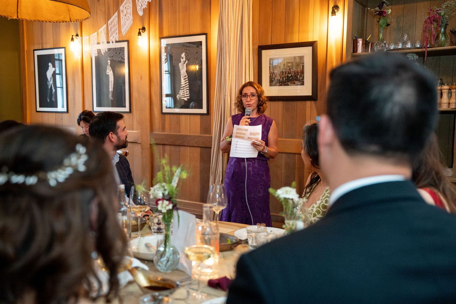 Woman in purple dress gives a speech at an elegant wedding reception dinner in a wood-paneled room.