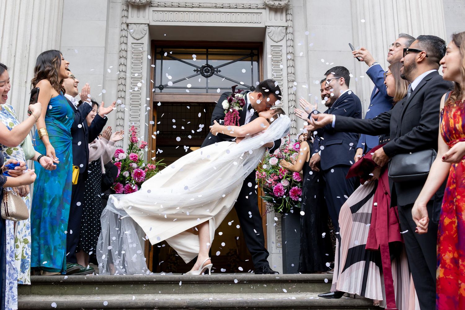 Bride and groom share a romantic dip kiss as guests throw confetti on grand building steps.