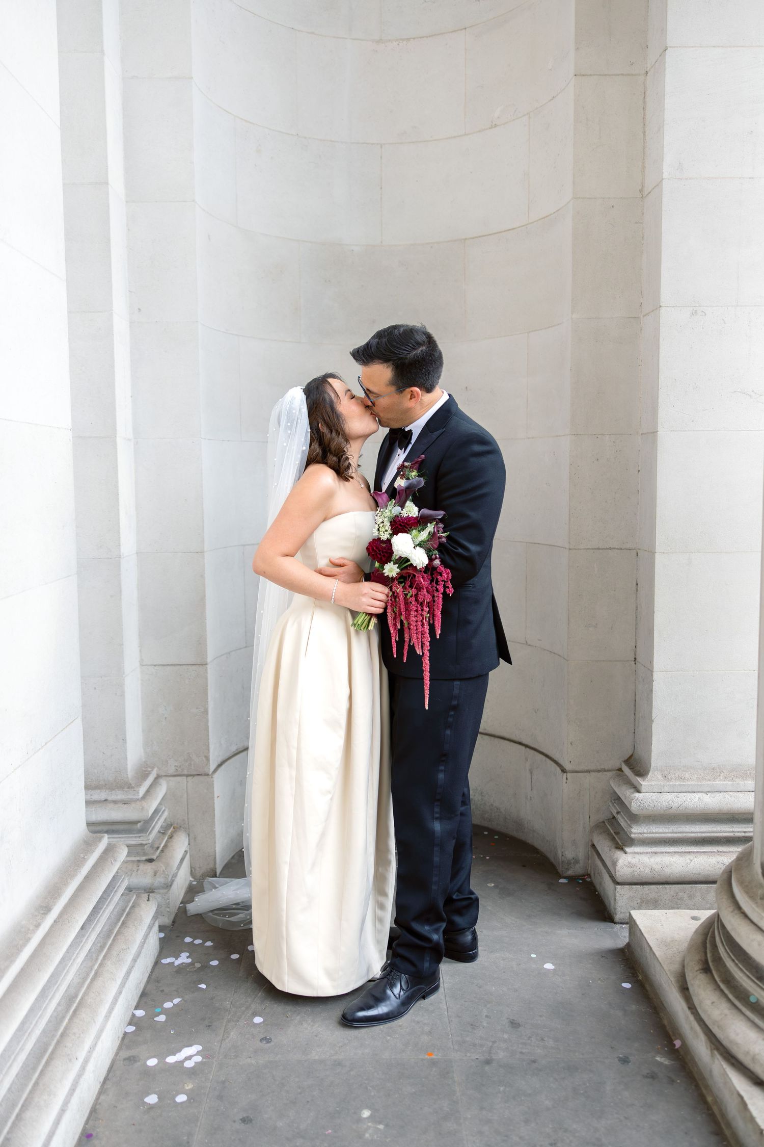 Bride and groom sharing first kiss outside stone building after London wedding ceremony.