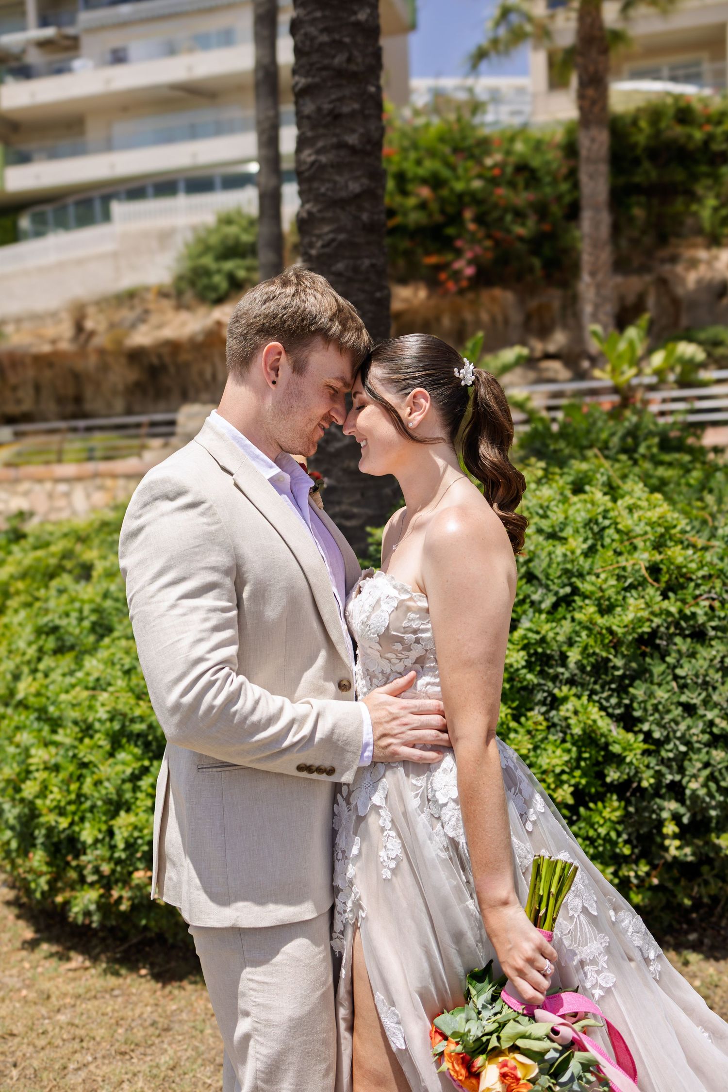 Romantic bride and groom touch foreheads tenderly in lush garden setting on sunny wedding day.