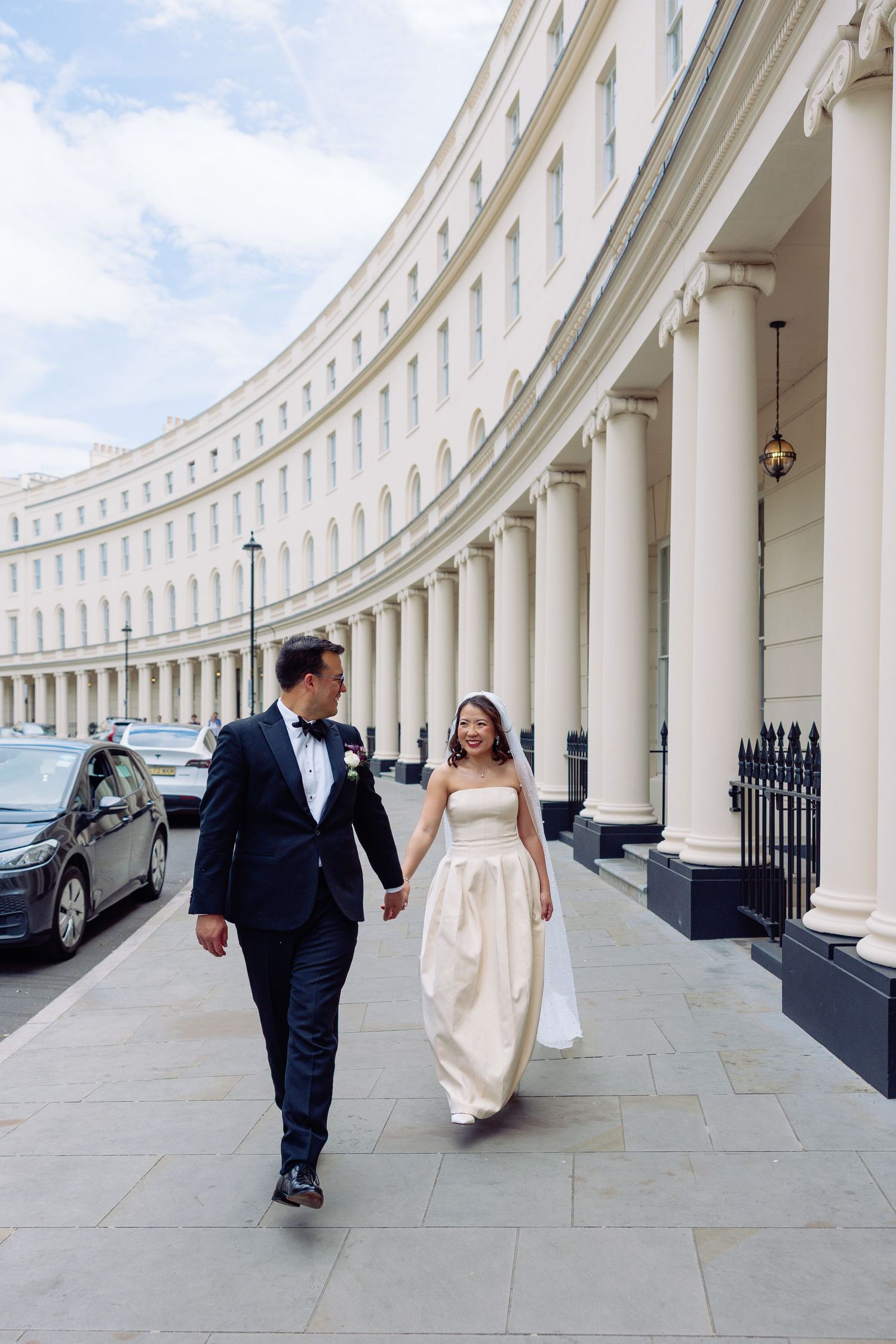 Happy newlyweds walk hand in hand along curved white Regency colonnade on wedding day.