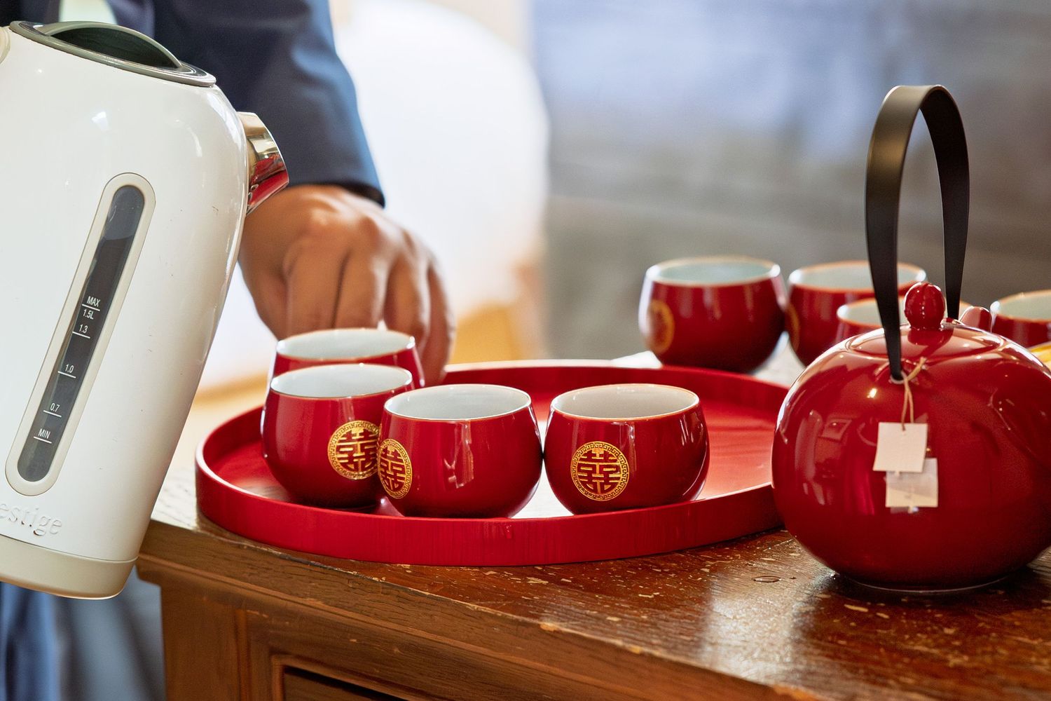 Red Chinese tea set with gold double happiness symbols on a wooden tray, being filled with hot water.
