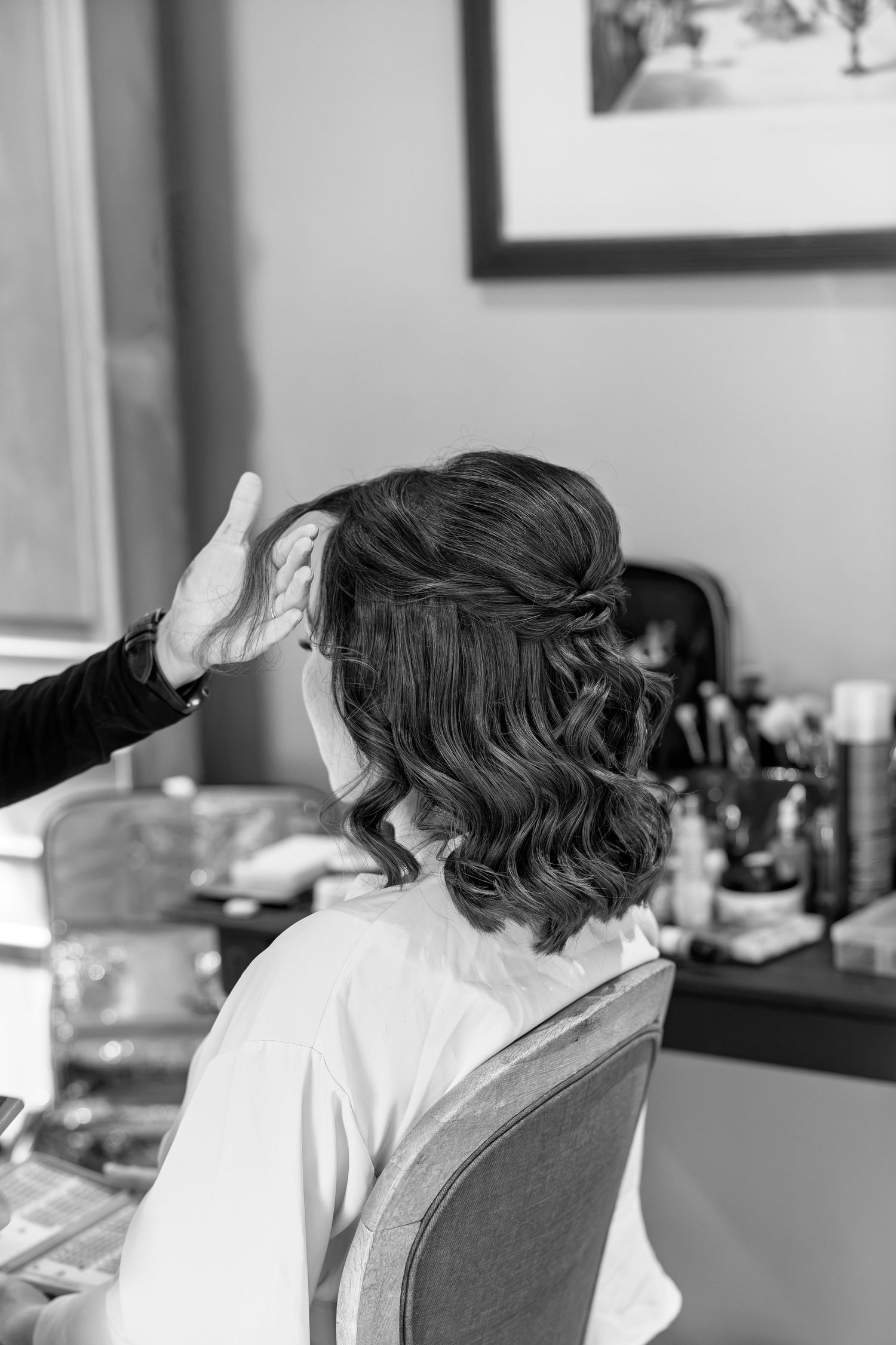 Hairstylist styling a bride's short wavy hair in a half-up do at a vanity mirror in black and white.