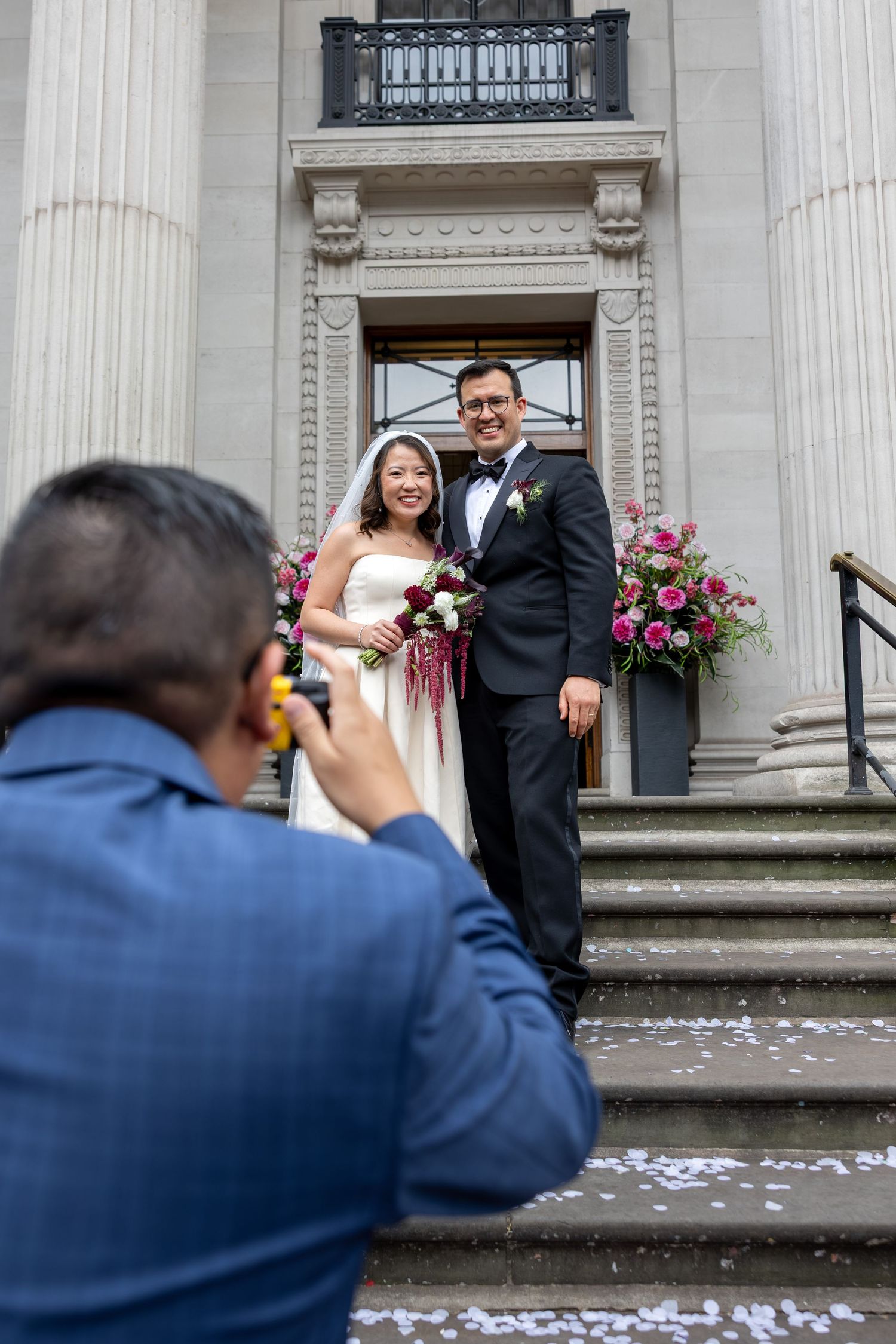 Guest photographs newlyweds posing on steps of neoclassical building with pink floral arrangements.