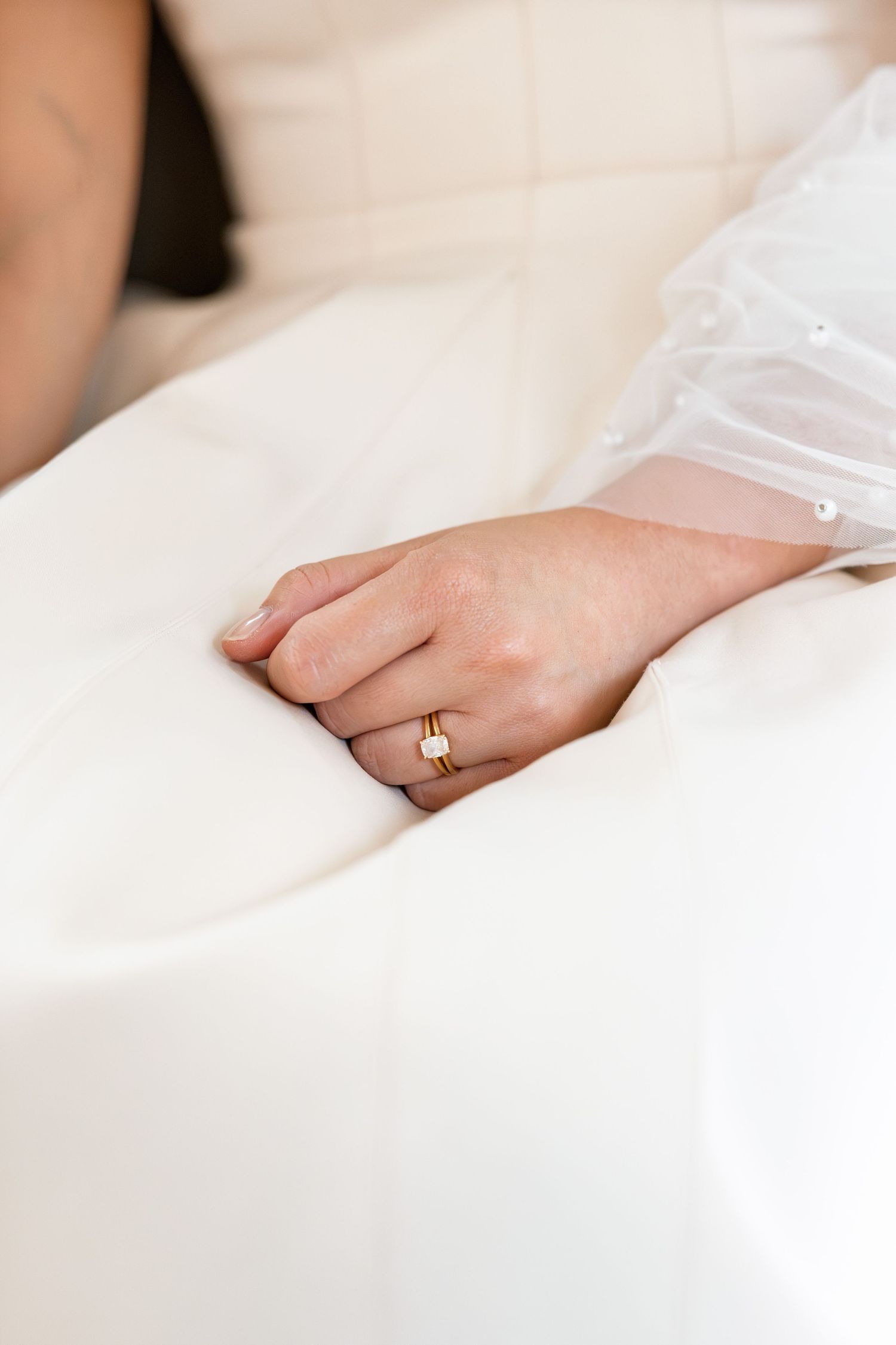 Close-up of a bride's hand resting on white wedding dress, wearing a gold diamond engagement ring.