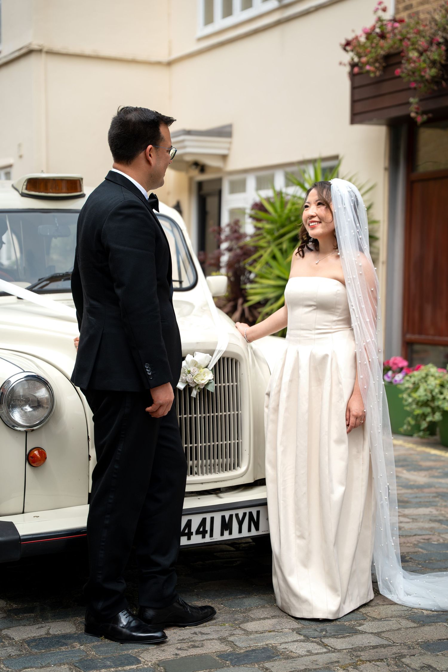 Bride in strapless white gown and veil with groom in tuxedo beside a vintage white wedding car.