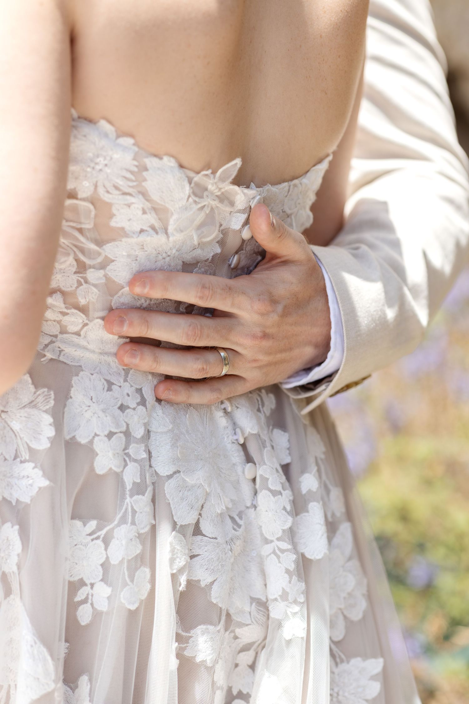 Close-up of groom's hand with wedding ring resting on bride's floral lace wedding dress back.