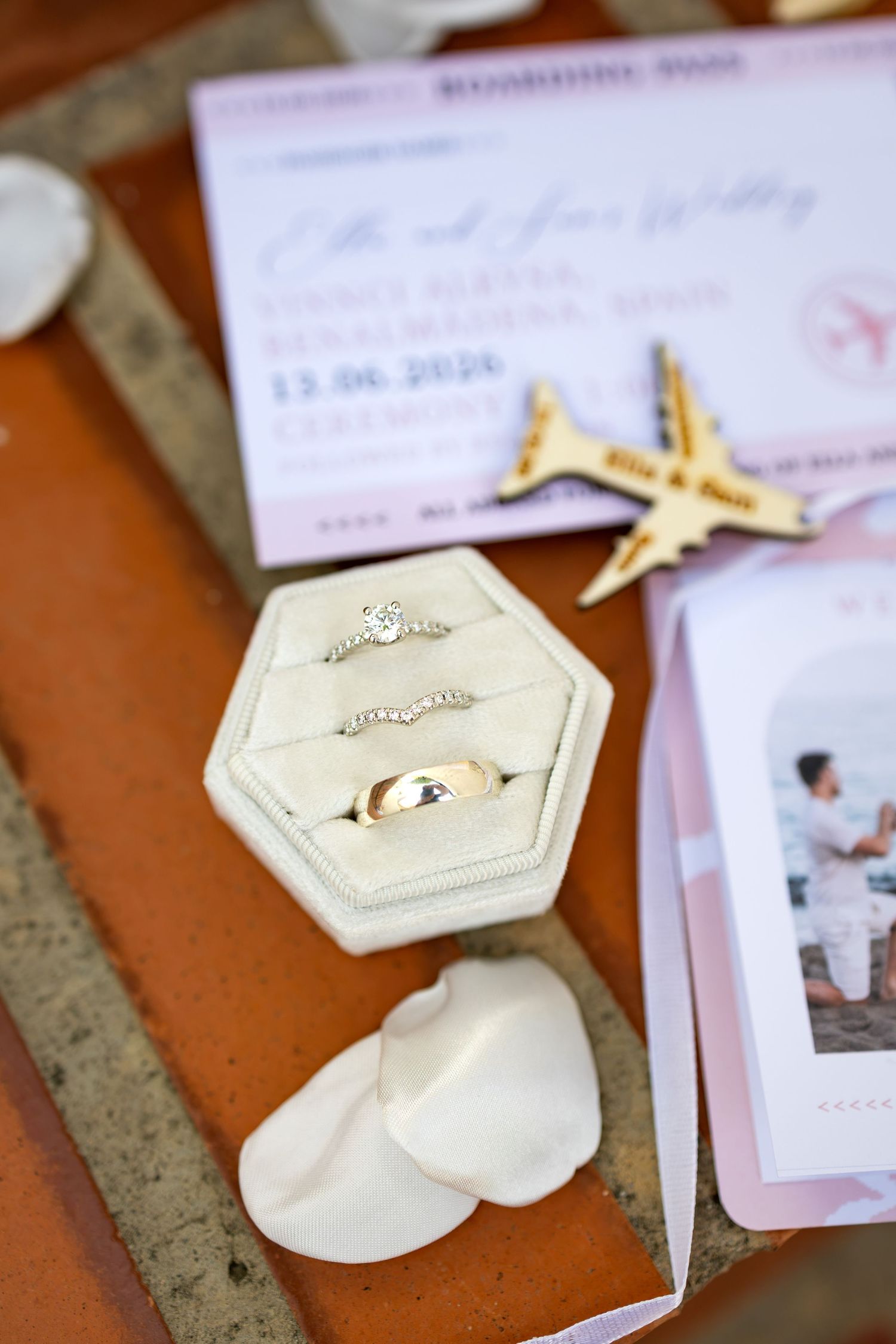Wedding rings in a velvet hexagon box with travel-themed invitations, a seashell, and a wooden airplane on terracotta tiles.