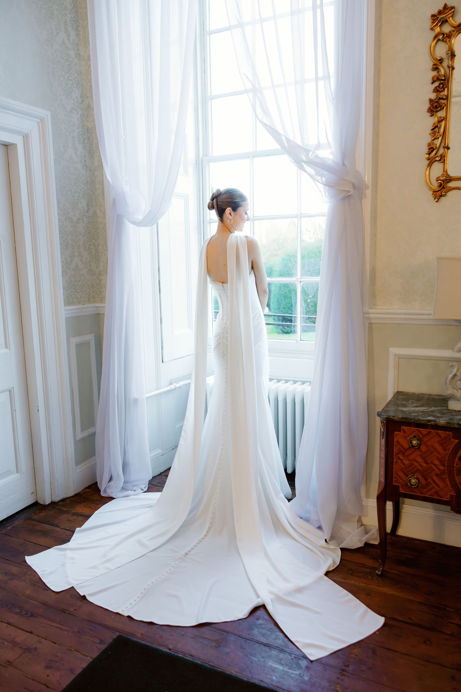 Elegant bridal portrait showing flowing train against white curtained window in vintage room.
