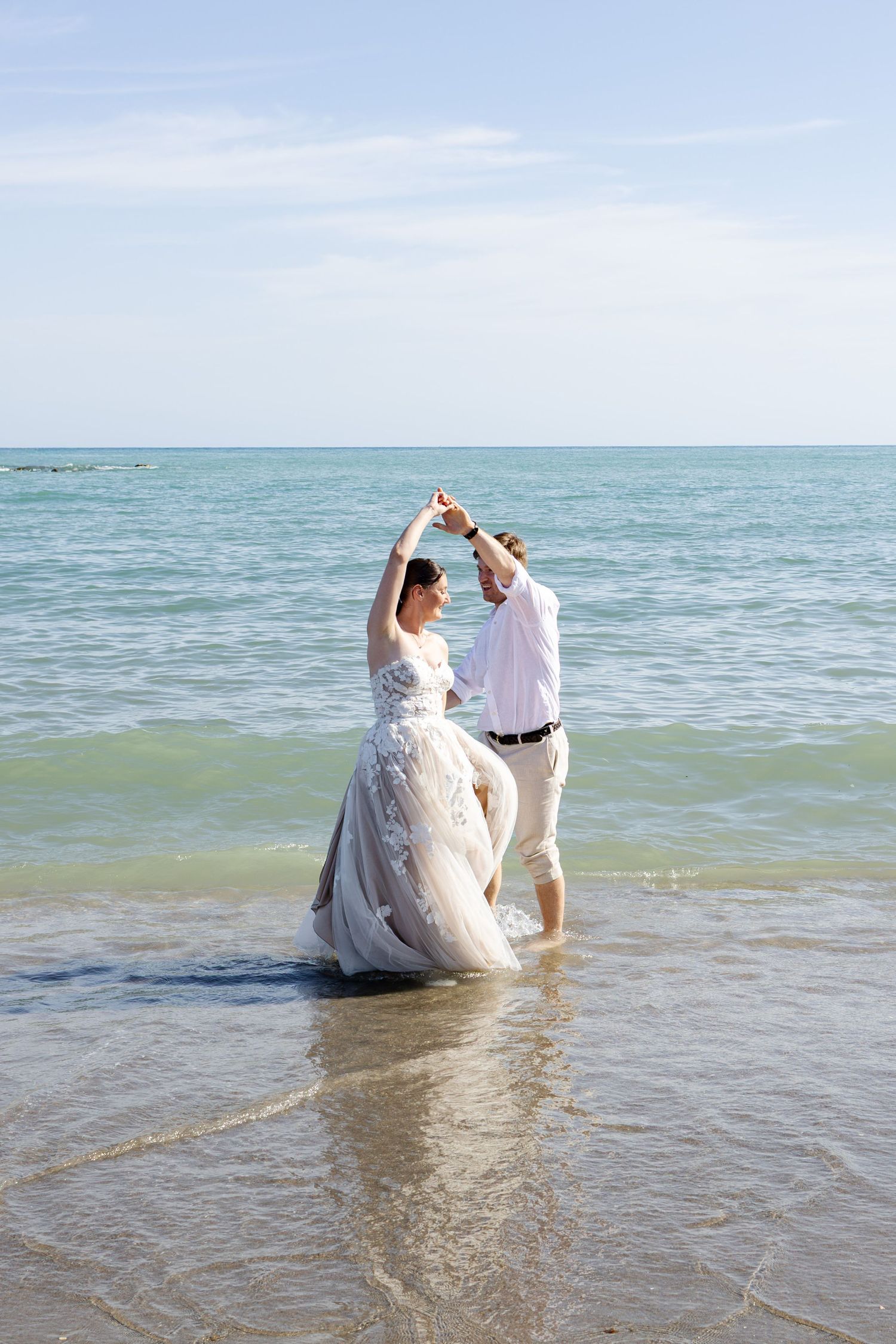 Bride and groom dance joyfully in ocean waves, arms raised, with turquoise sea and blue sky behind them.