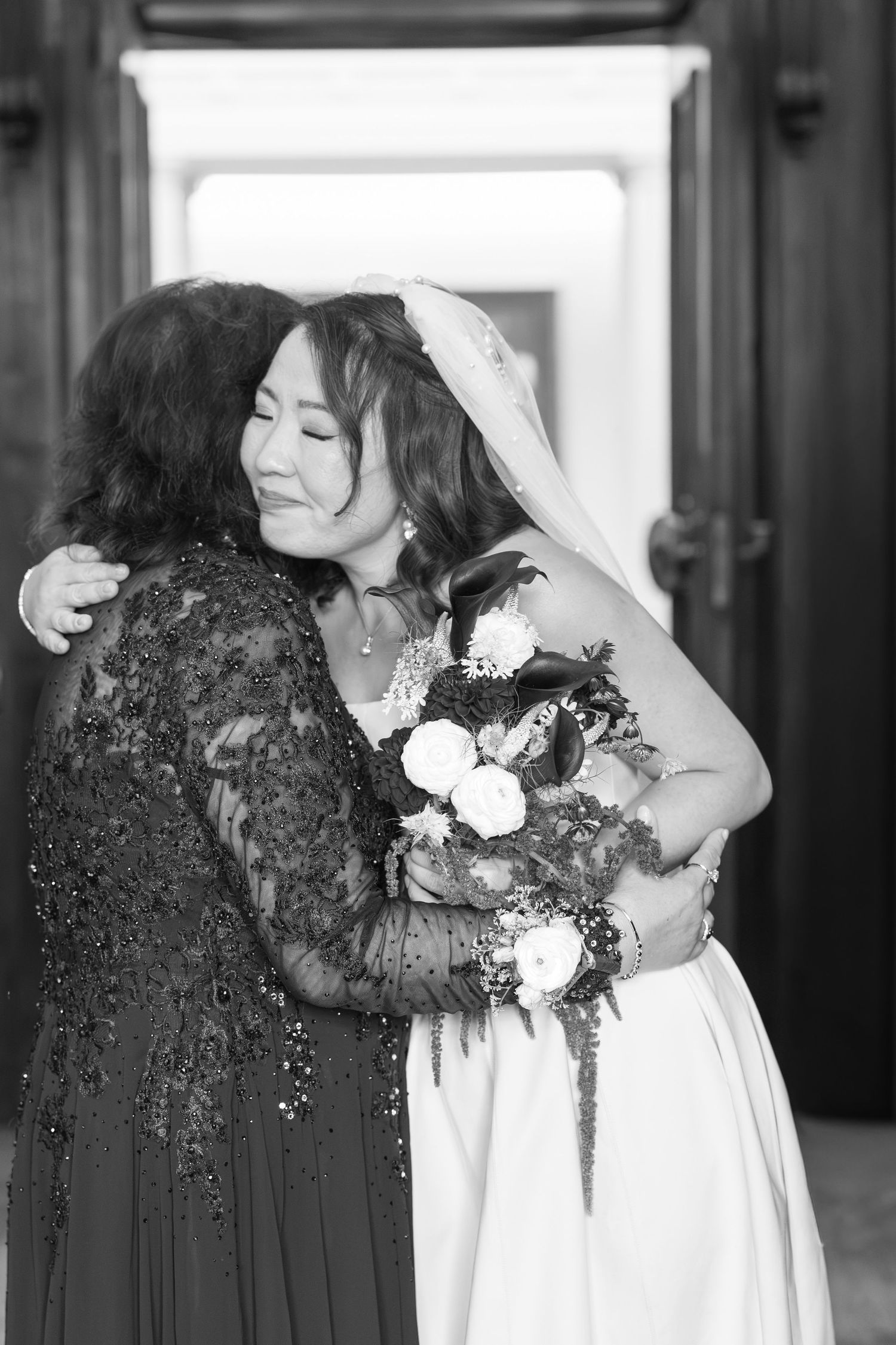 Black and white photo of a bride in a veil hugging an older woman in lace dress while holding a bouquet.