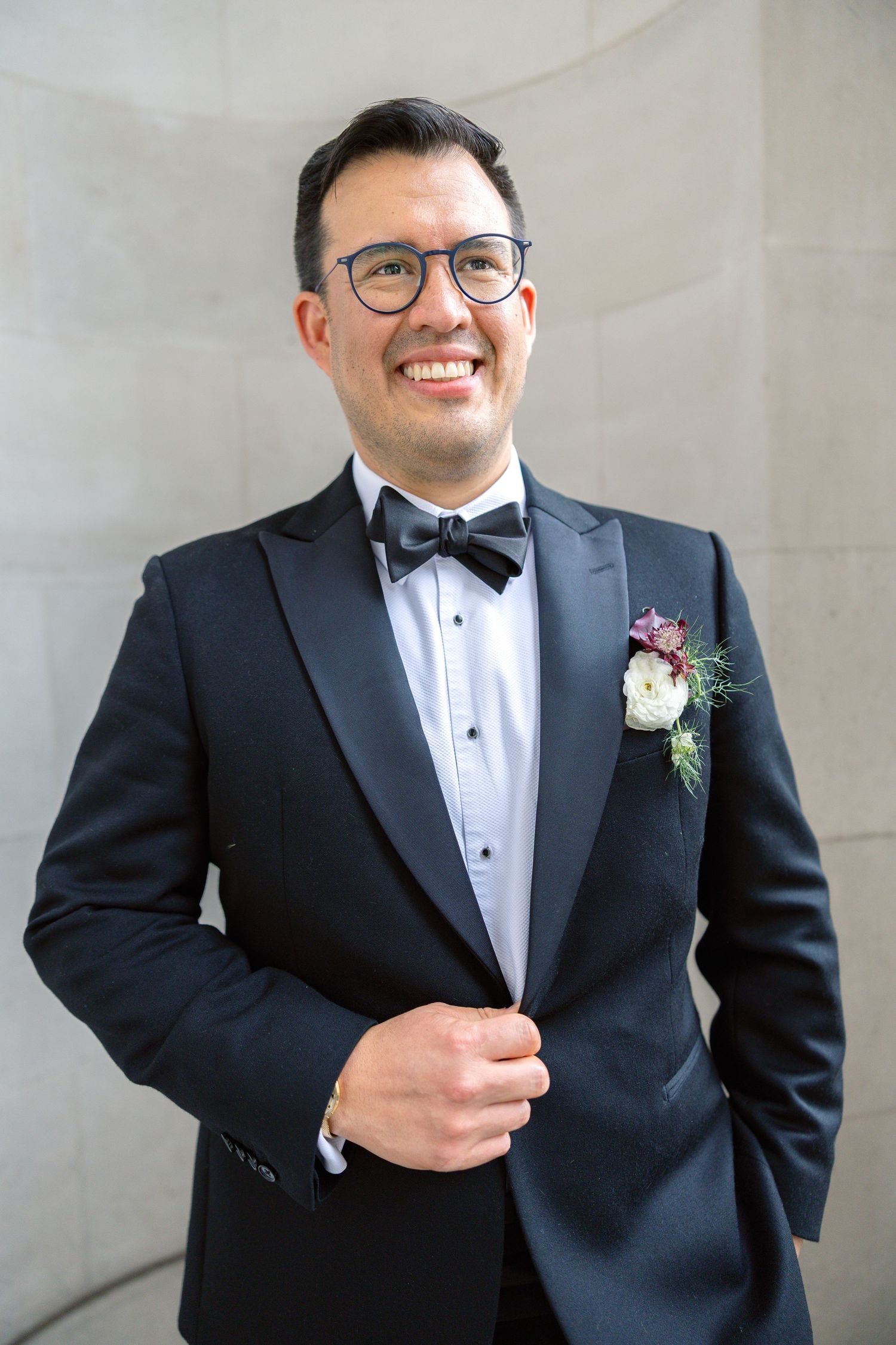 Smiling groom in black tuxedo with boutonniere and bow tie wearing glasses.