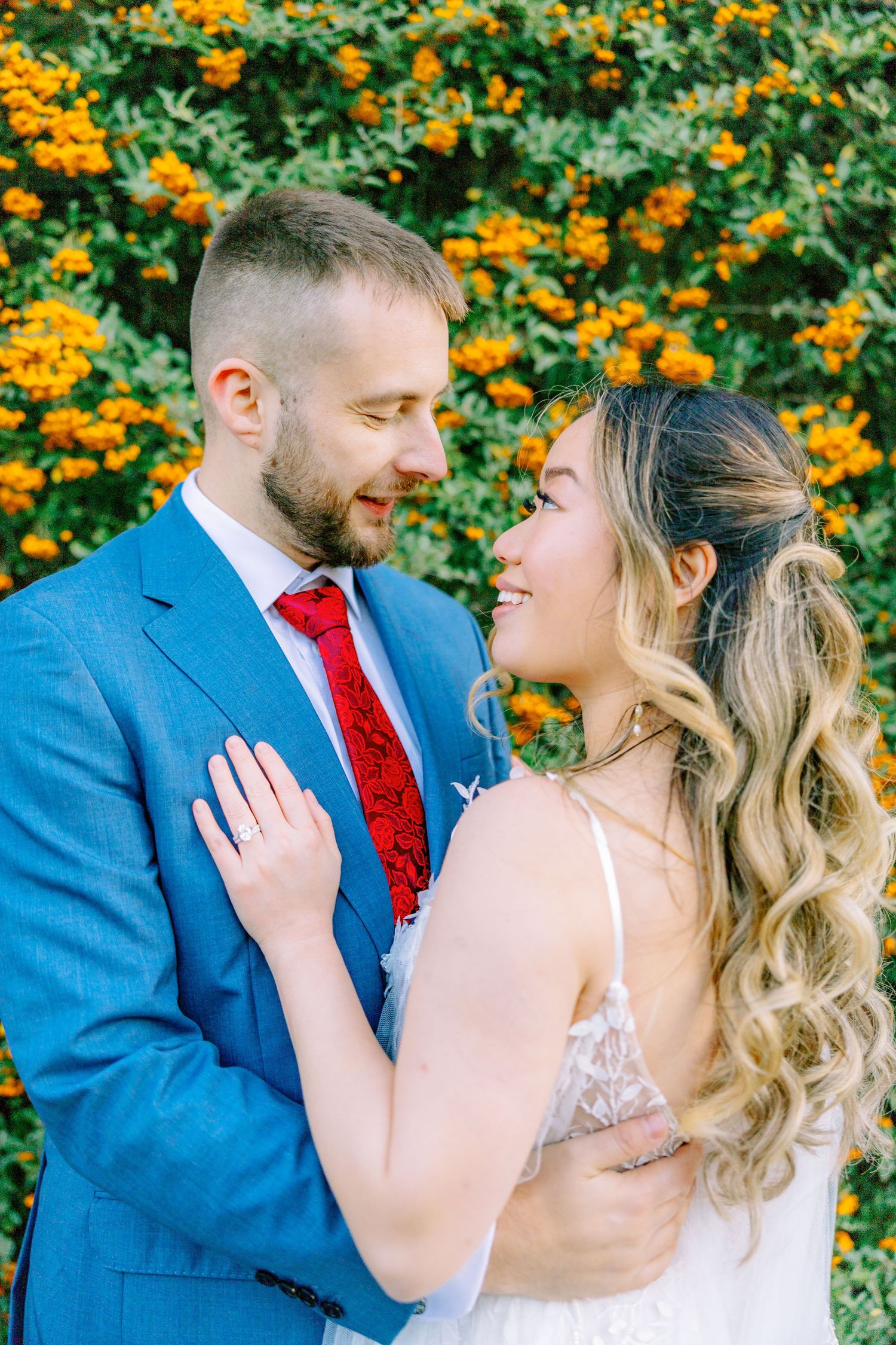 Wedding couple poses in front of vibrant orange marigolds, with the groom in blue suit and red tie.