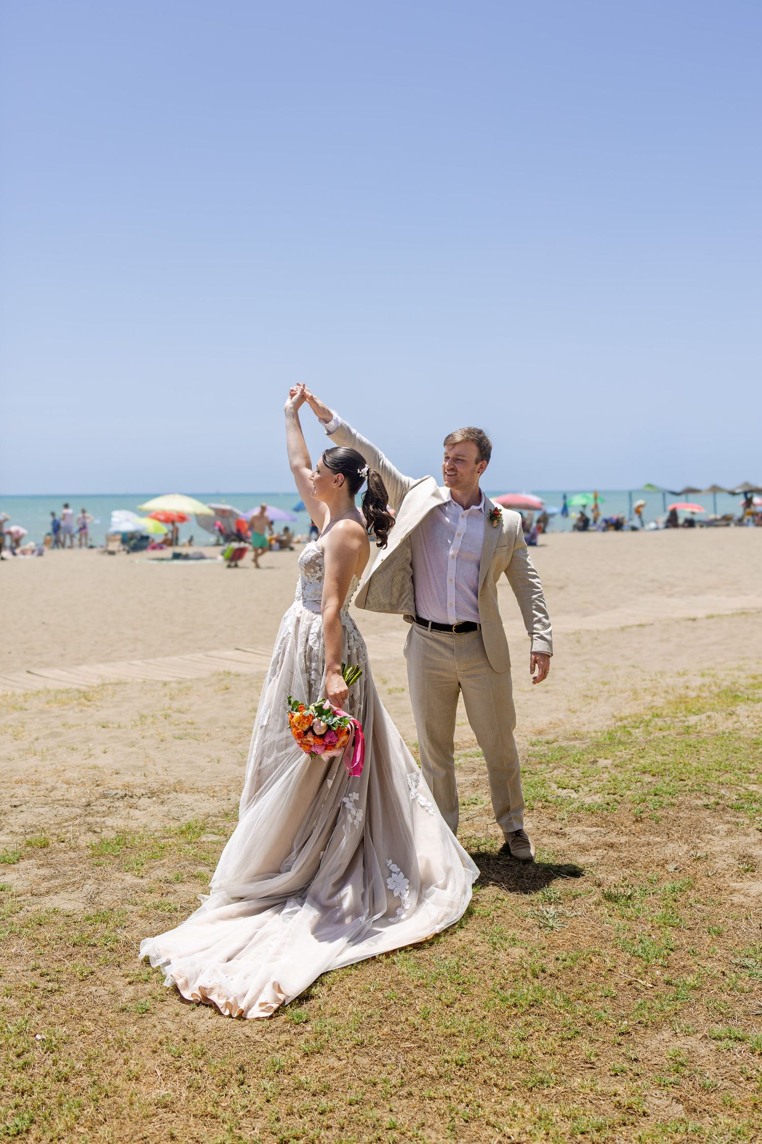 Bride and groom dancing on a sunny beach, bride twirling in a floral gown holding a colorful bouquet.