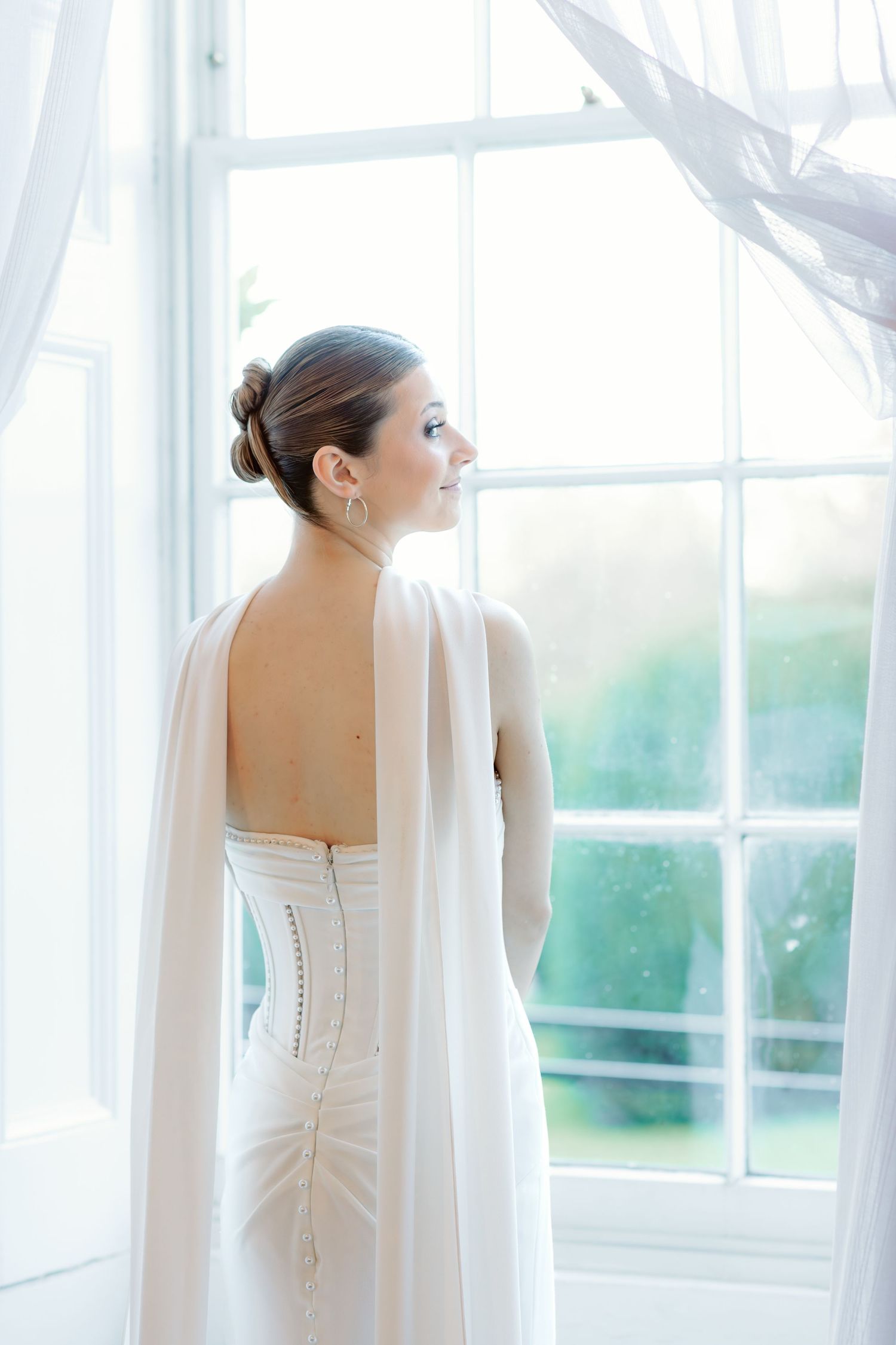 Back view of wedding dress with sheer cape by sunlit window in elegant room.