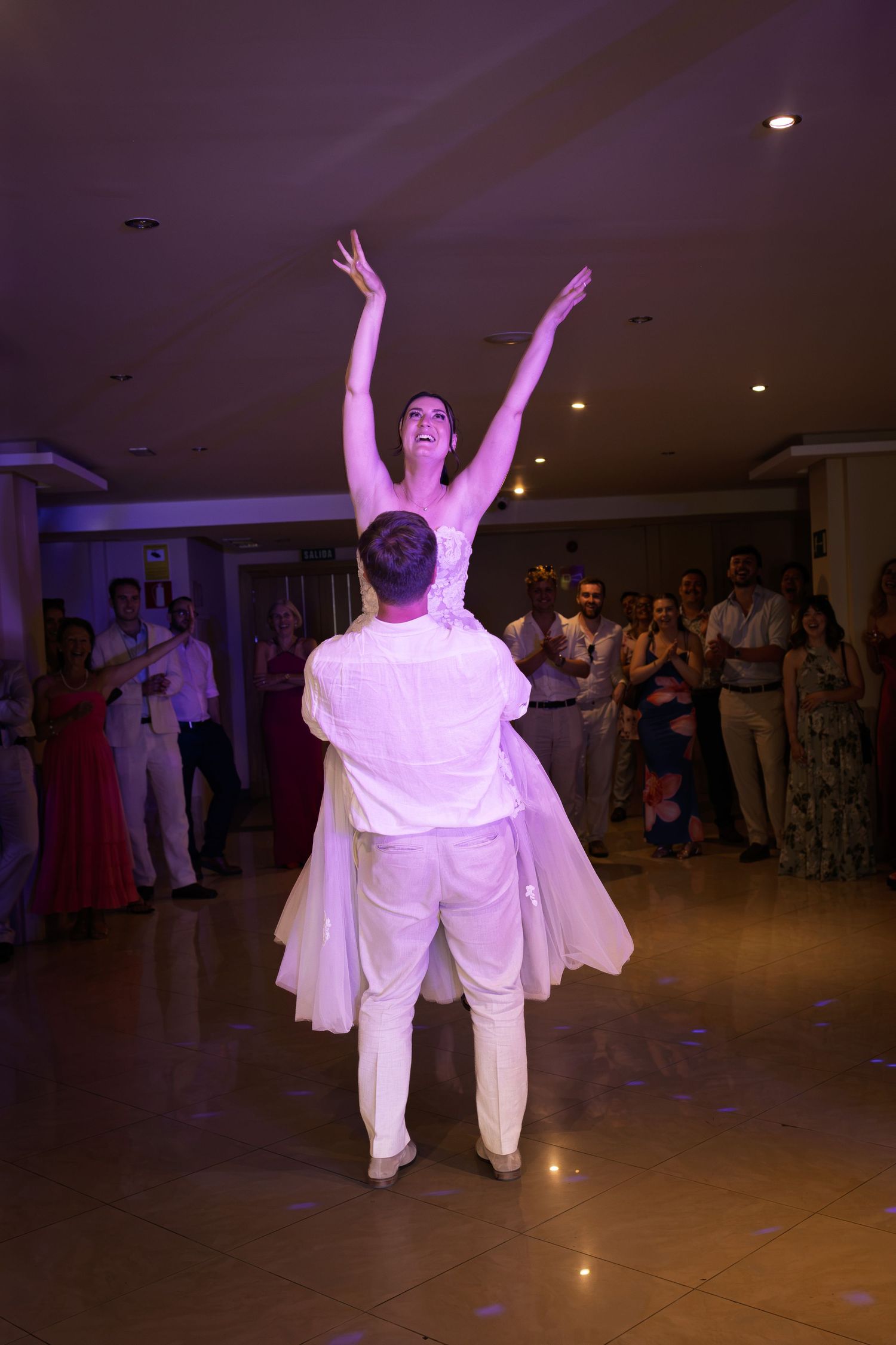 Groom lifts radiant bride with arms raised joyfully during energetic first dance at wedding reception.