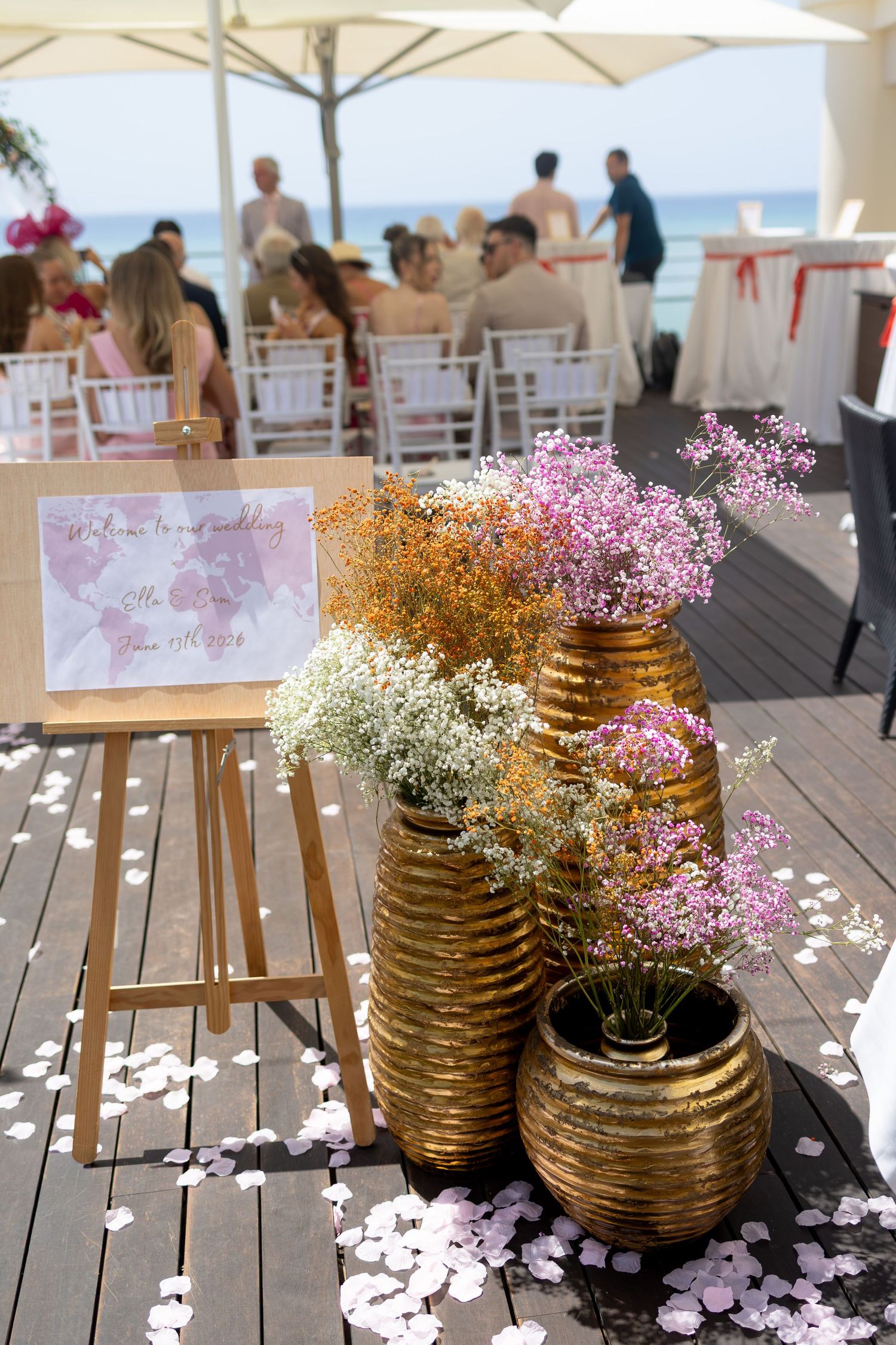 Golden vases with colorful flowers decorate an outdoor beachside wedding reception with white chairs and guests.
