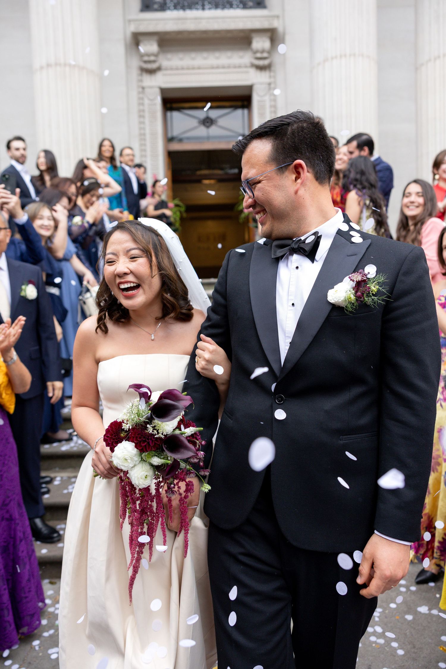Joyful newlyweds exit ceremony venue through shower of white confetti thrown by guests.