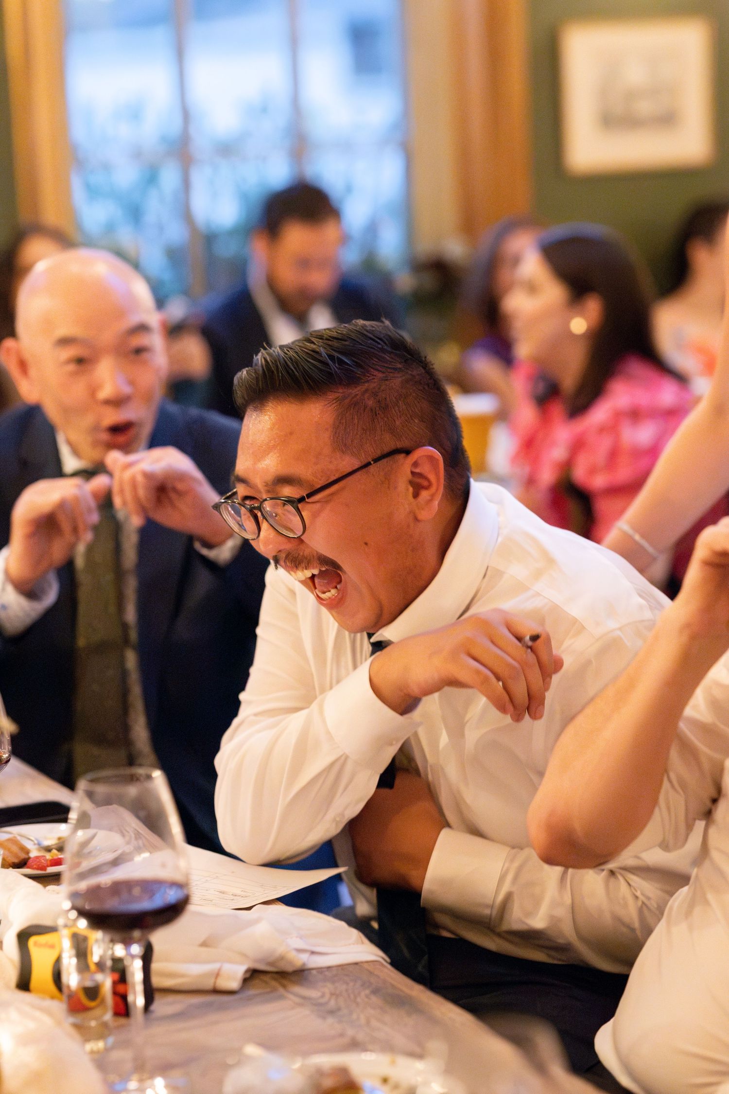 Excited wedding guest reacts enthusiastically at lively reception dinner table with wine glasses.