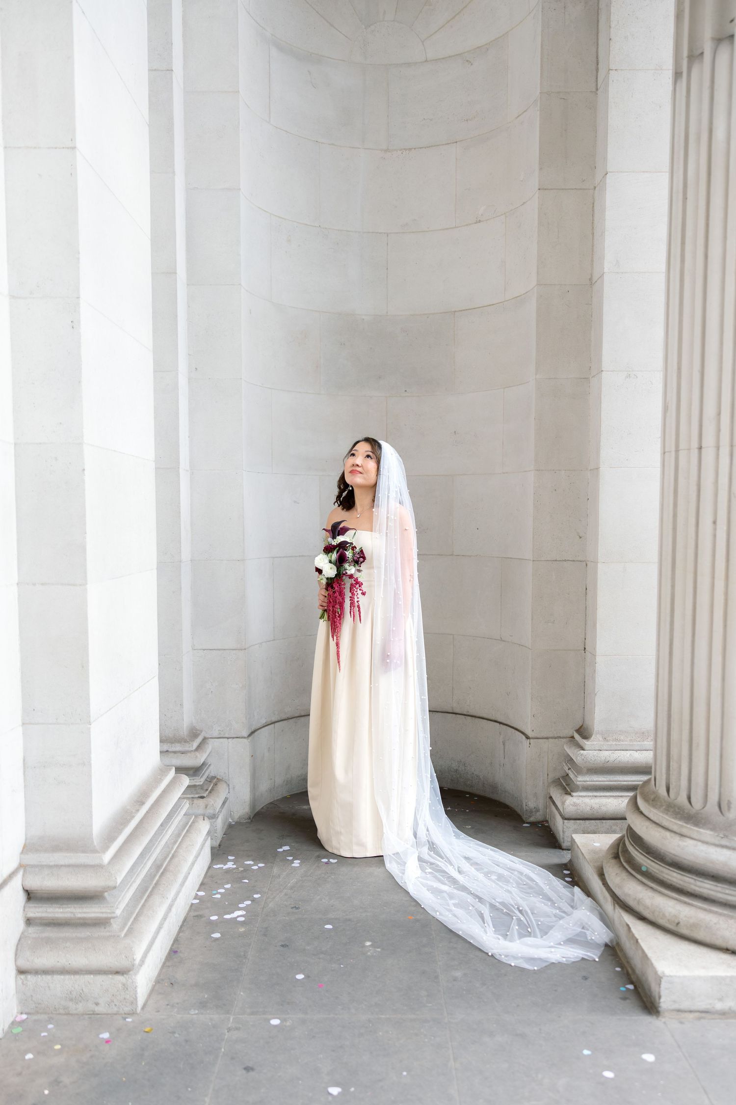 Bride in strapless ivory gown with flowing cathedral veil gazes upward between stone pillars.