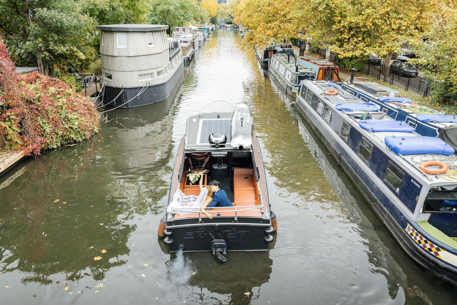 Canal boats line the waterway as newlyweds pose for photos on their vessel.