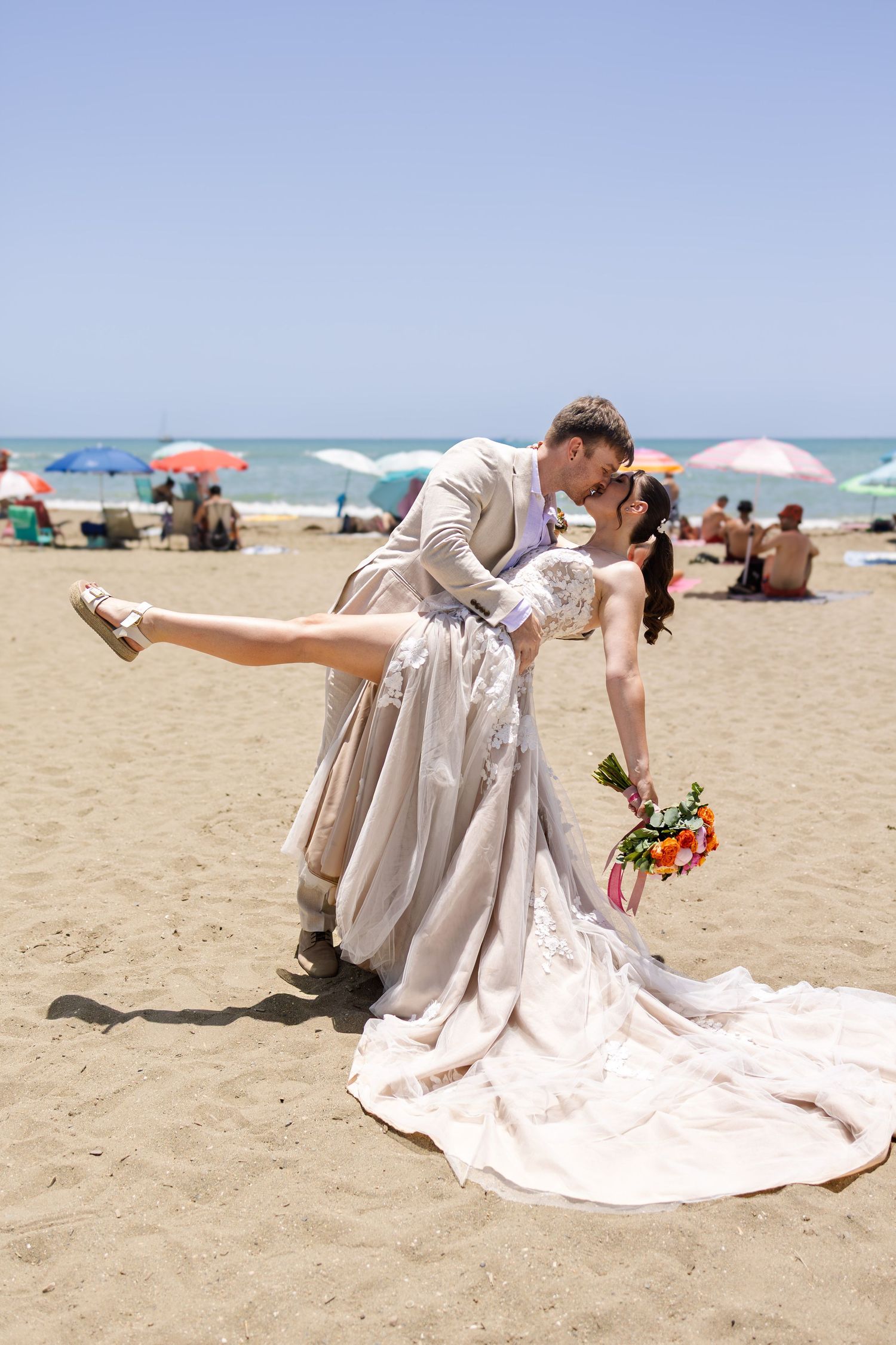 Groom dipping bride for a romantic kiss on a sunny beach with colorful umbrellas in background.