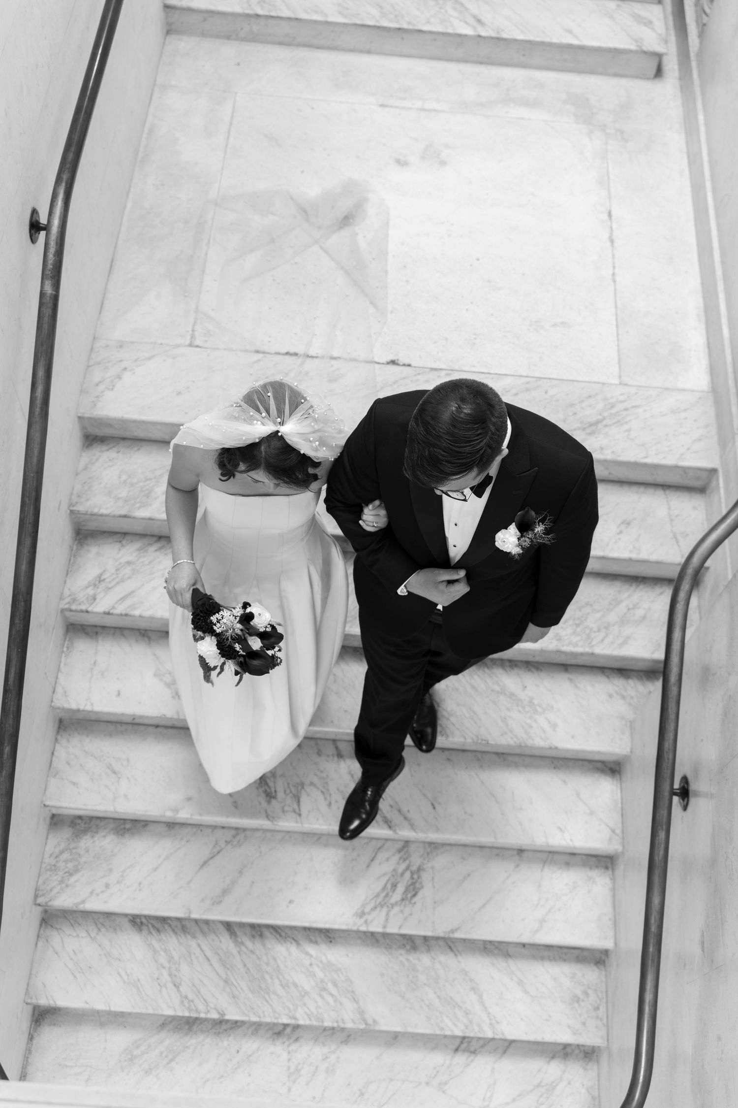 Aerial black and white photo of bride and groom walking arm in arm down marble staircase at wedding.