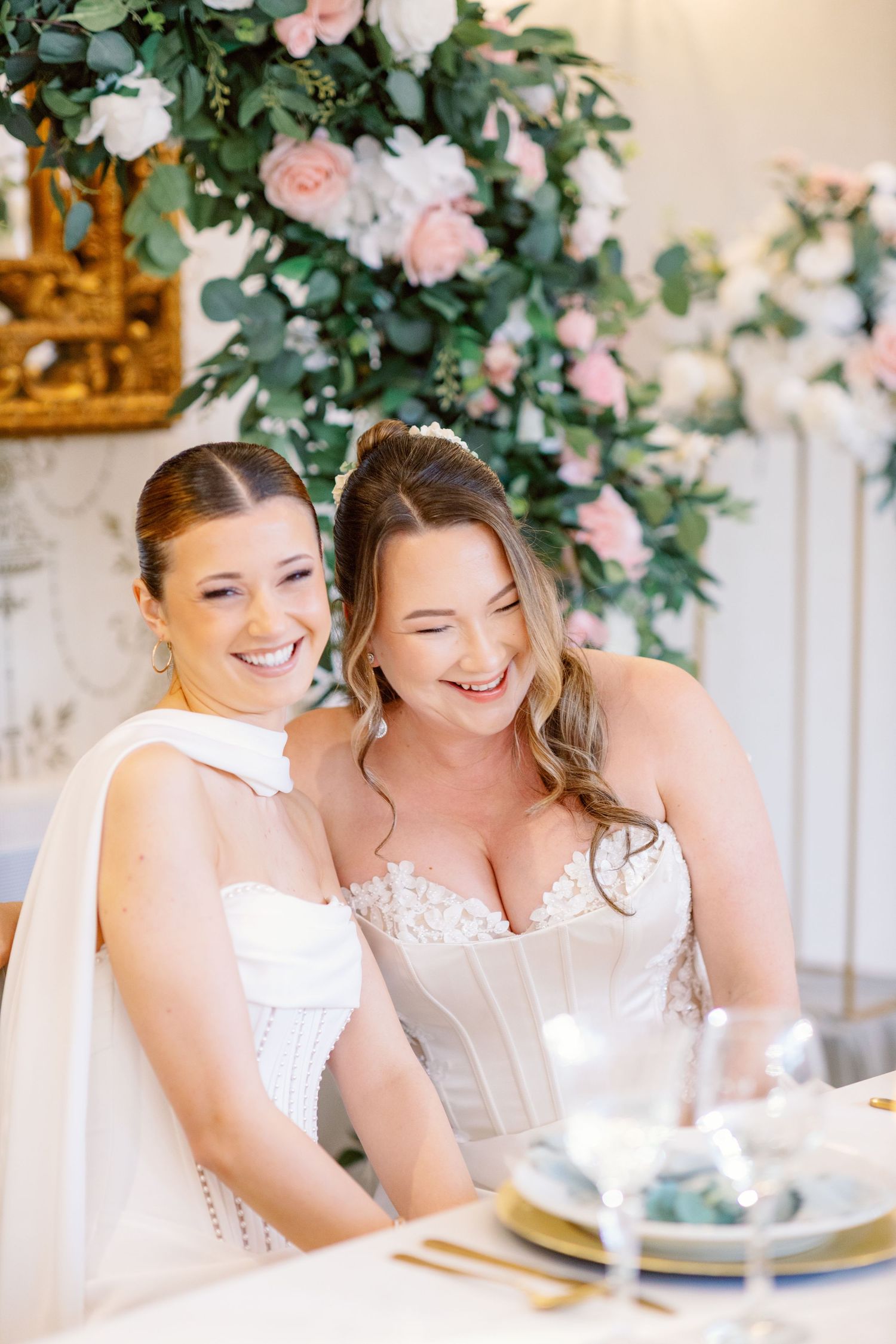 Two people in bridal attire share laughter while seated at an elegantly decorated table with floral decor.
