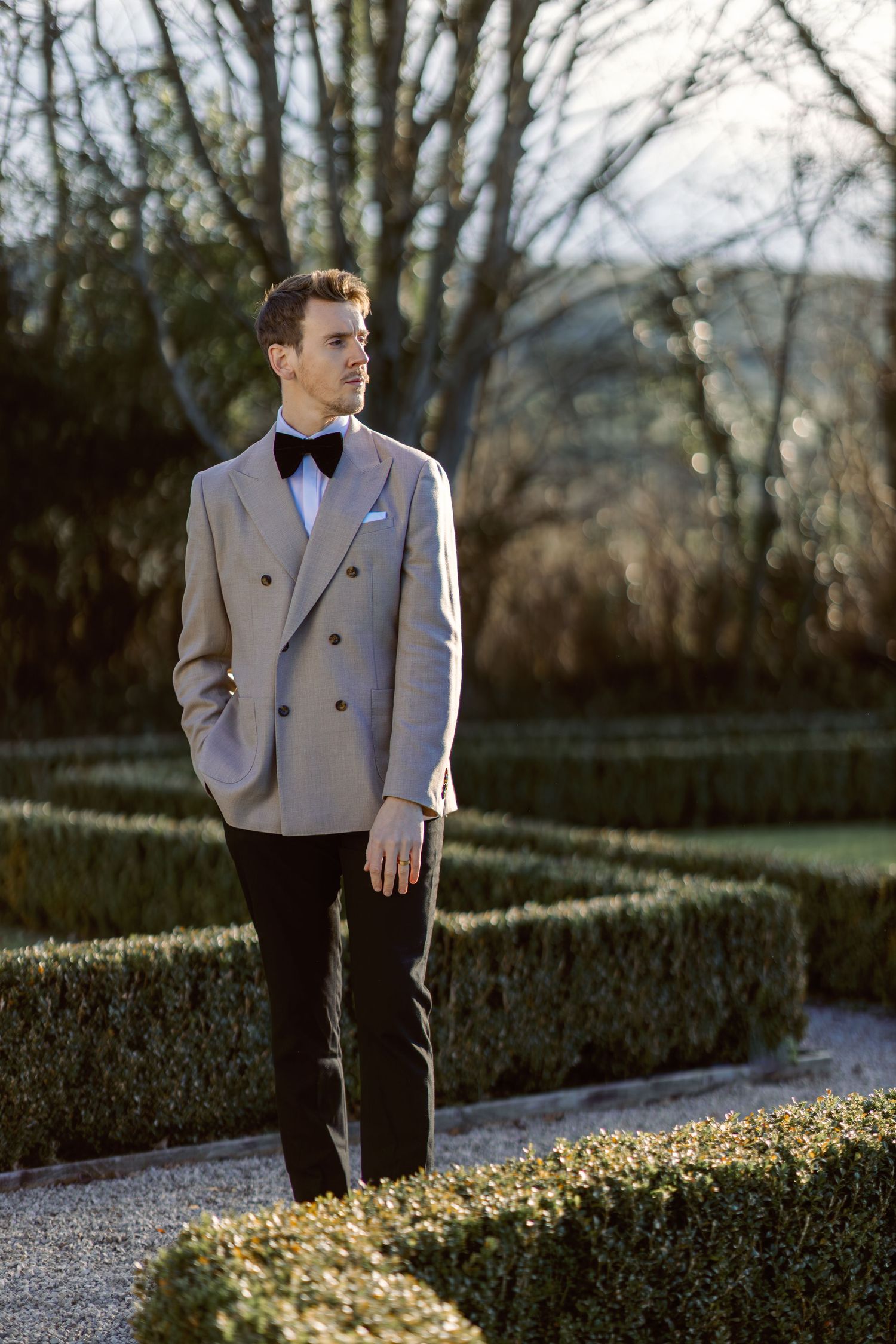Elegant groom in light colored suit jacket and bowtie standing in manicured garden path.