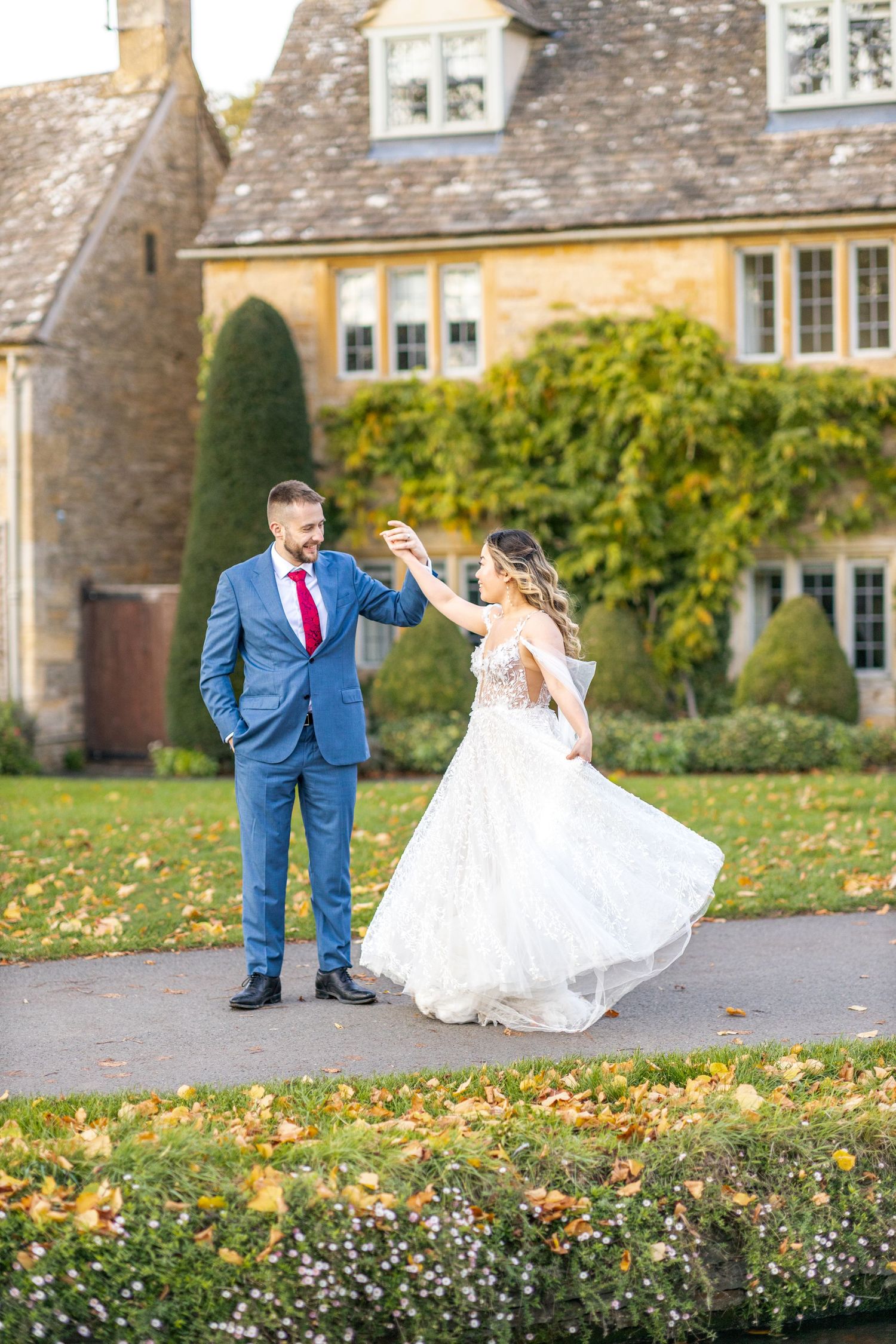 A couple in wedding attire playfully poses on fallen autumn leaves near a stone building.