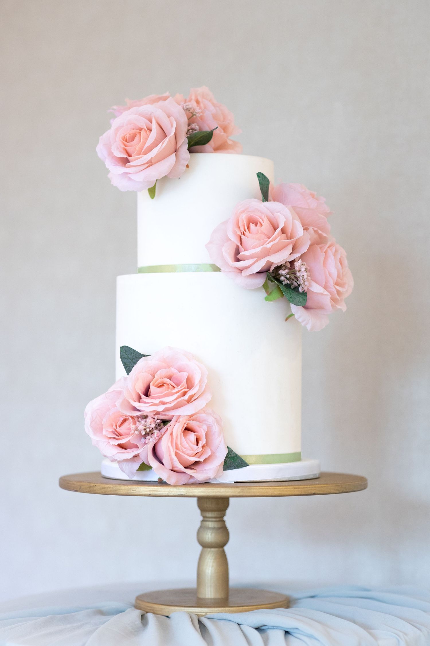 Elegant white wedding cake decorated with pink roses on a gold cake stand against a light gray background.