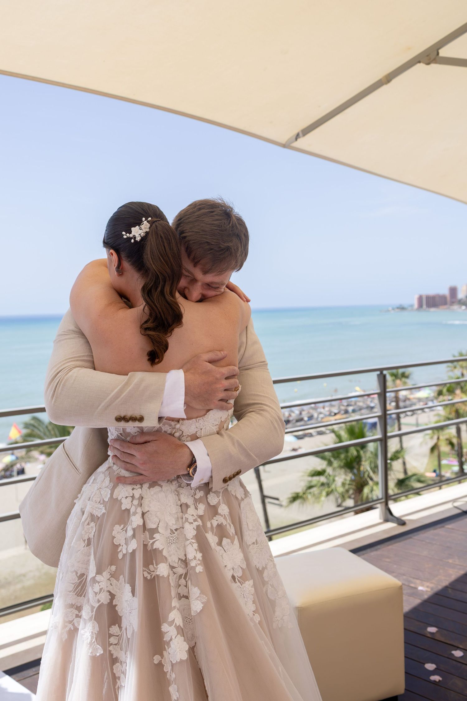 Bride and groom share tender embrace on seaside terrace, bride wearing floral lace wedding gown.