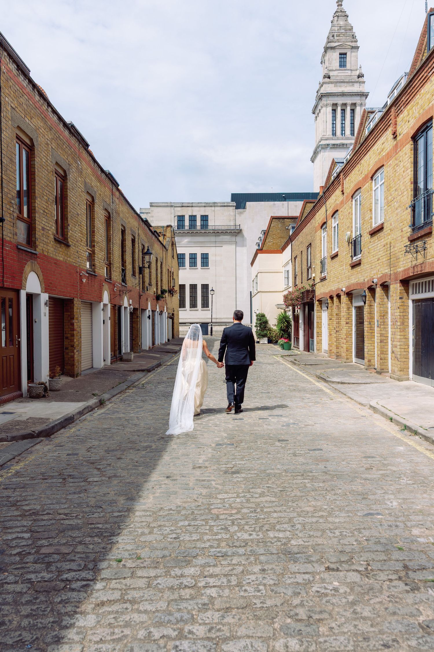 A bride and groom walk hand-in-hand down a cobblestone street lined with brick buildings and a church tower.