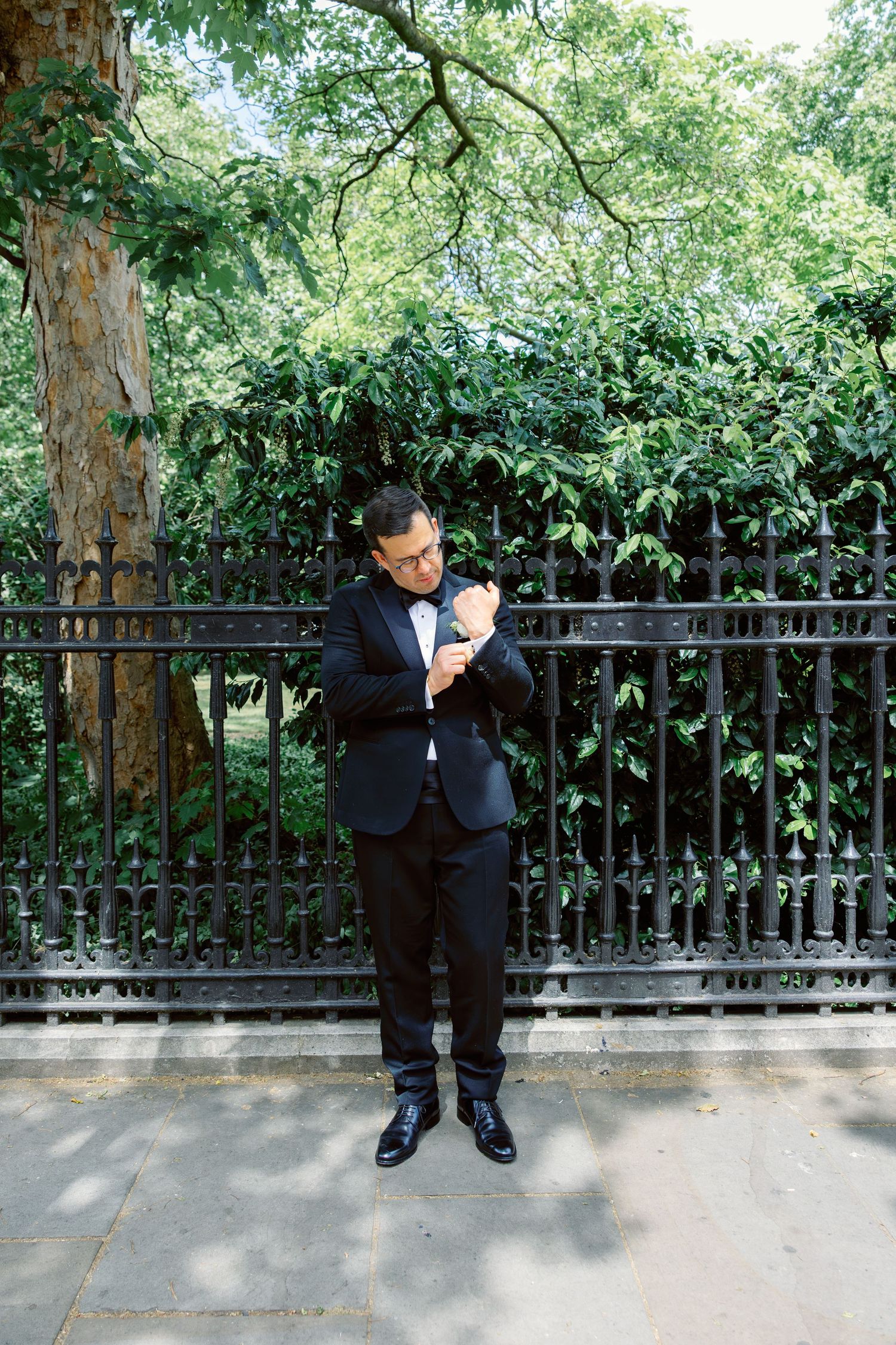 Groom in black tuxedo adjusting cufflinks outdoors near iron fence and trees.