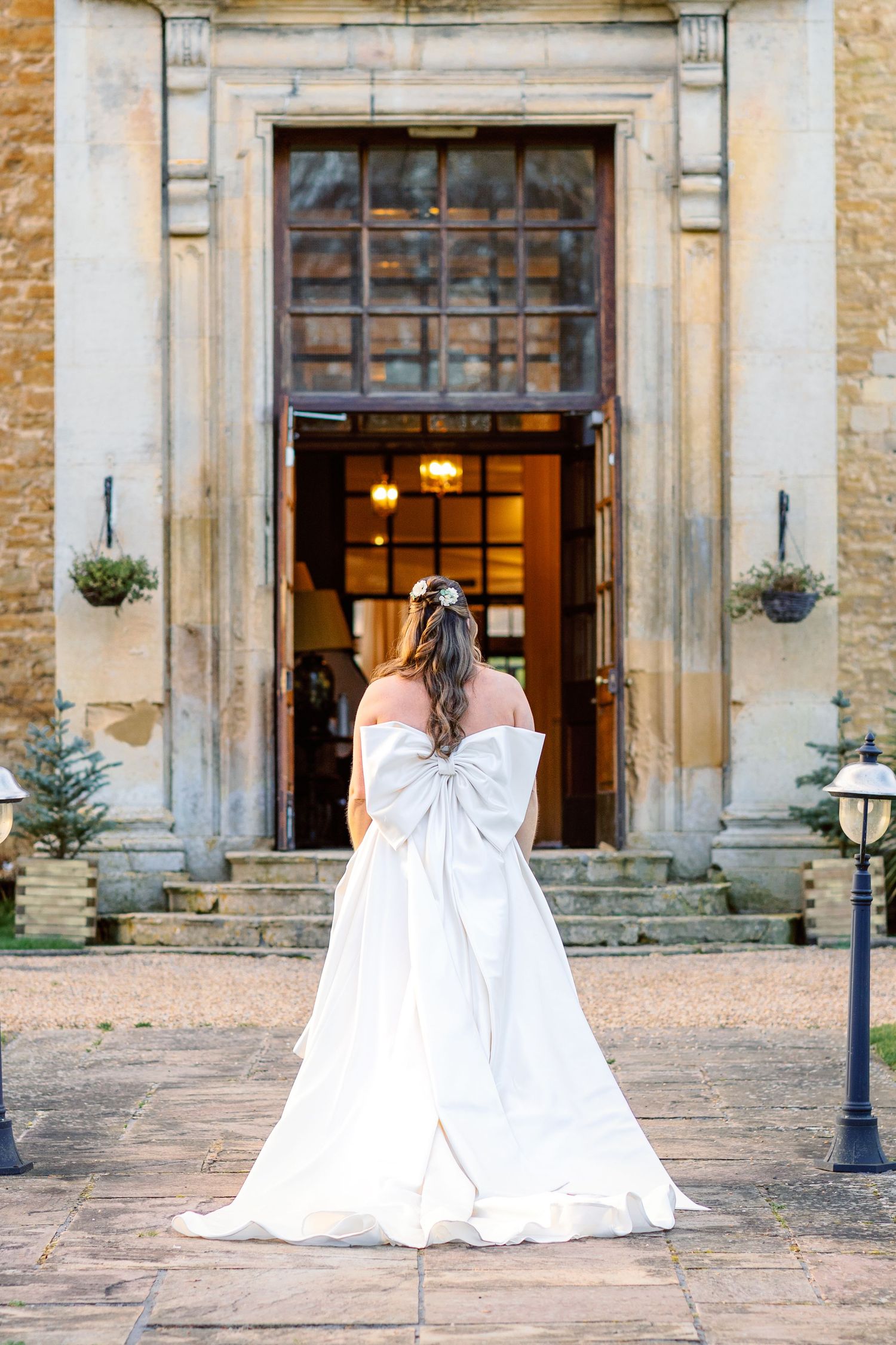 A bride in a flowing white wedding gown stands before grand entrance doors of a historic stone building.