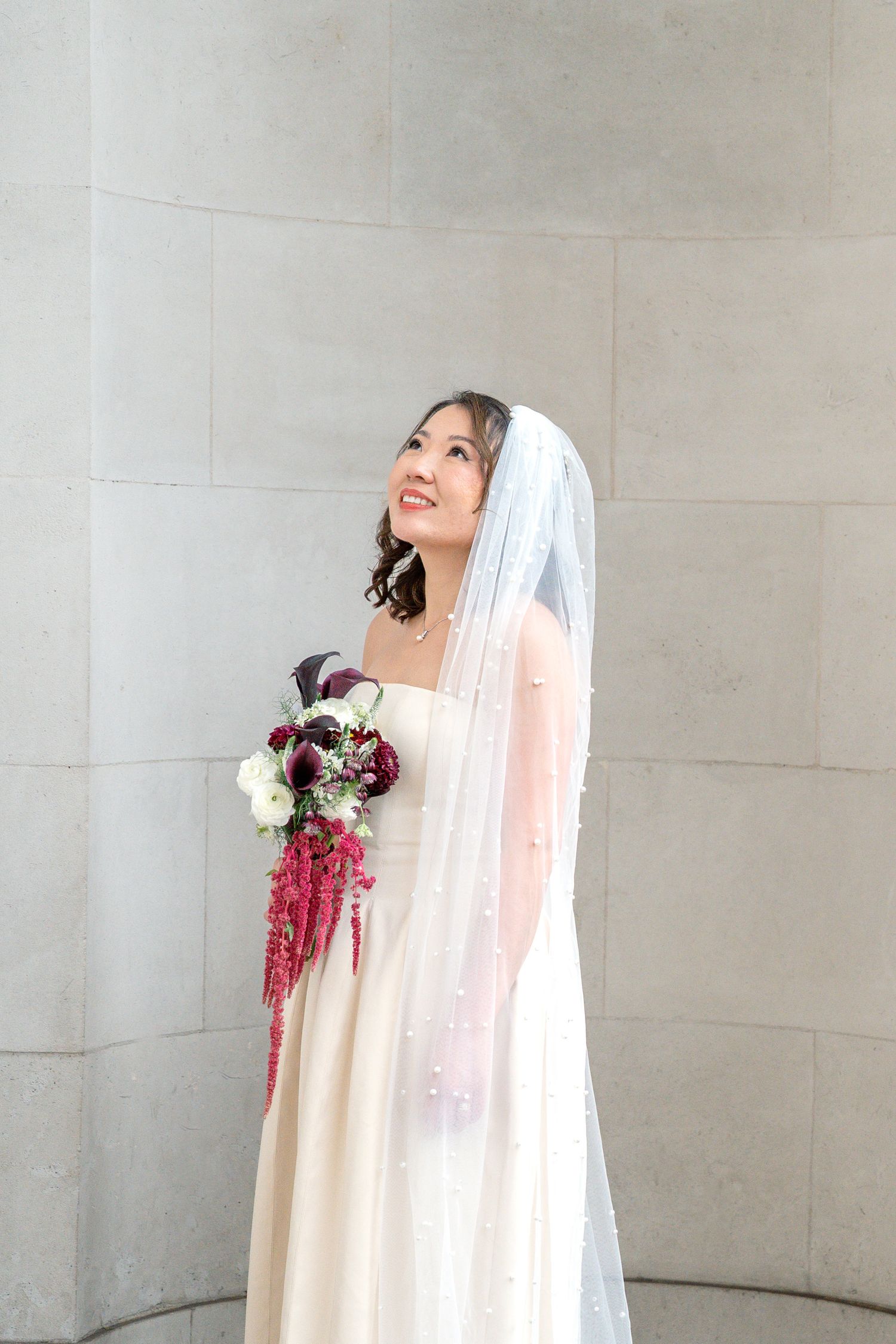 A bride in a white wedding gown and pearl-adorned veil holds a burgundy and white bouquet by stone columns.