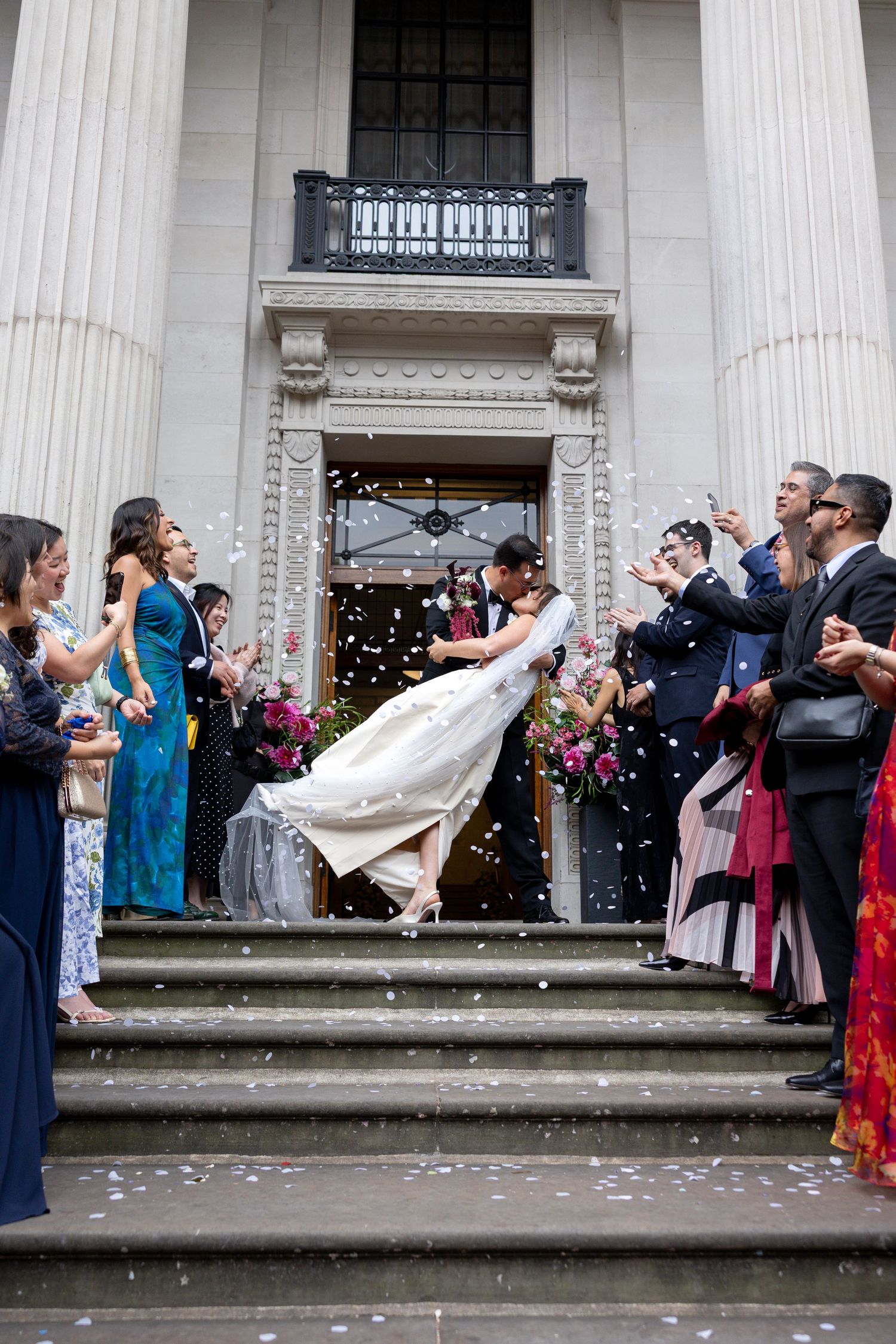 Groom dips bride on church steps while guests toss confetti at outdoor wedding ceremony exit.