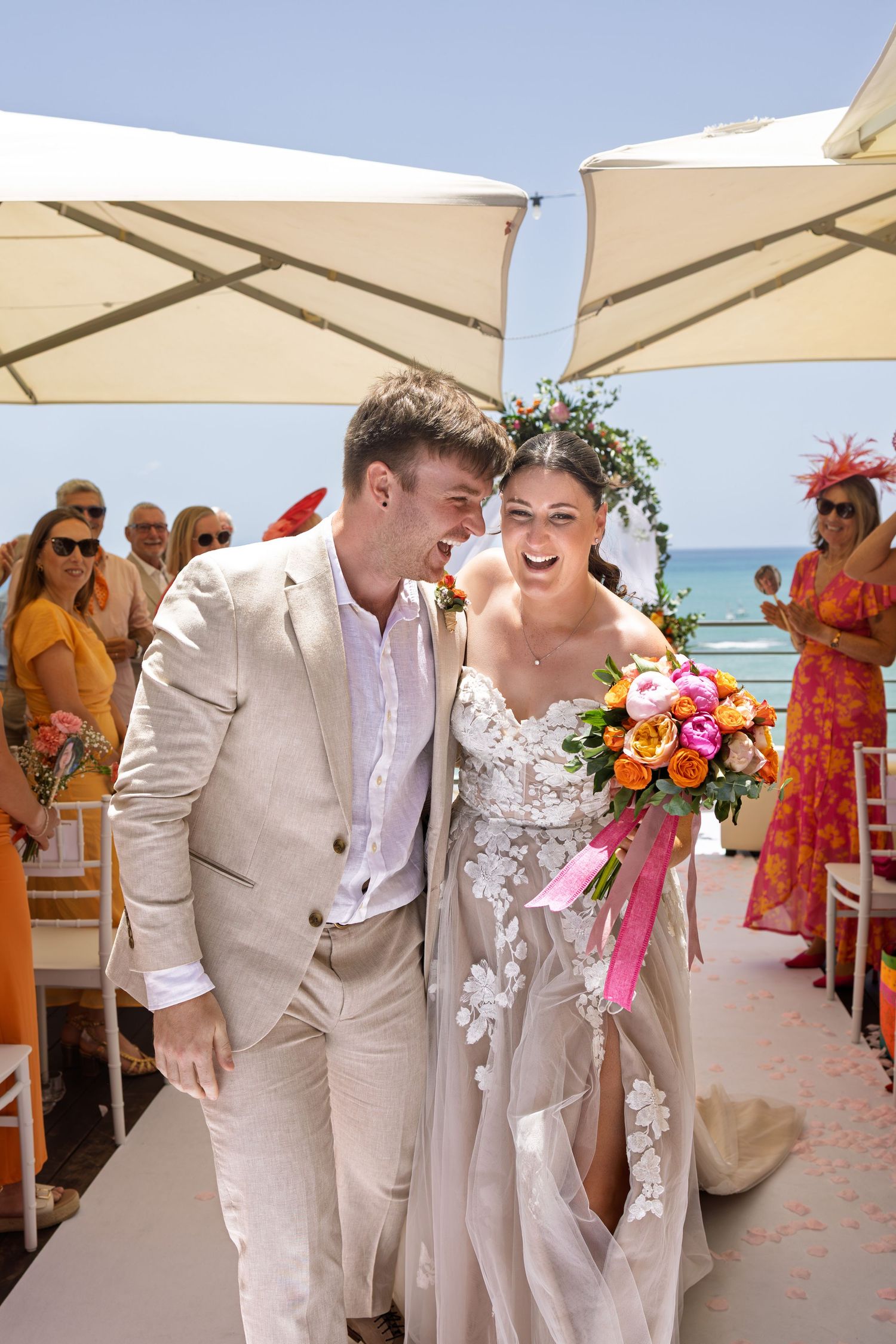 Joyful bride and groom walking down the aisle at a beachside wedding ceremony, bride holding colorful bouquet.