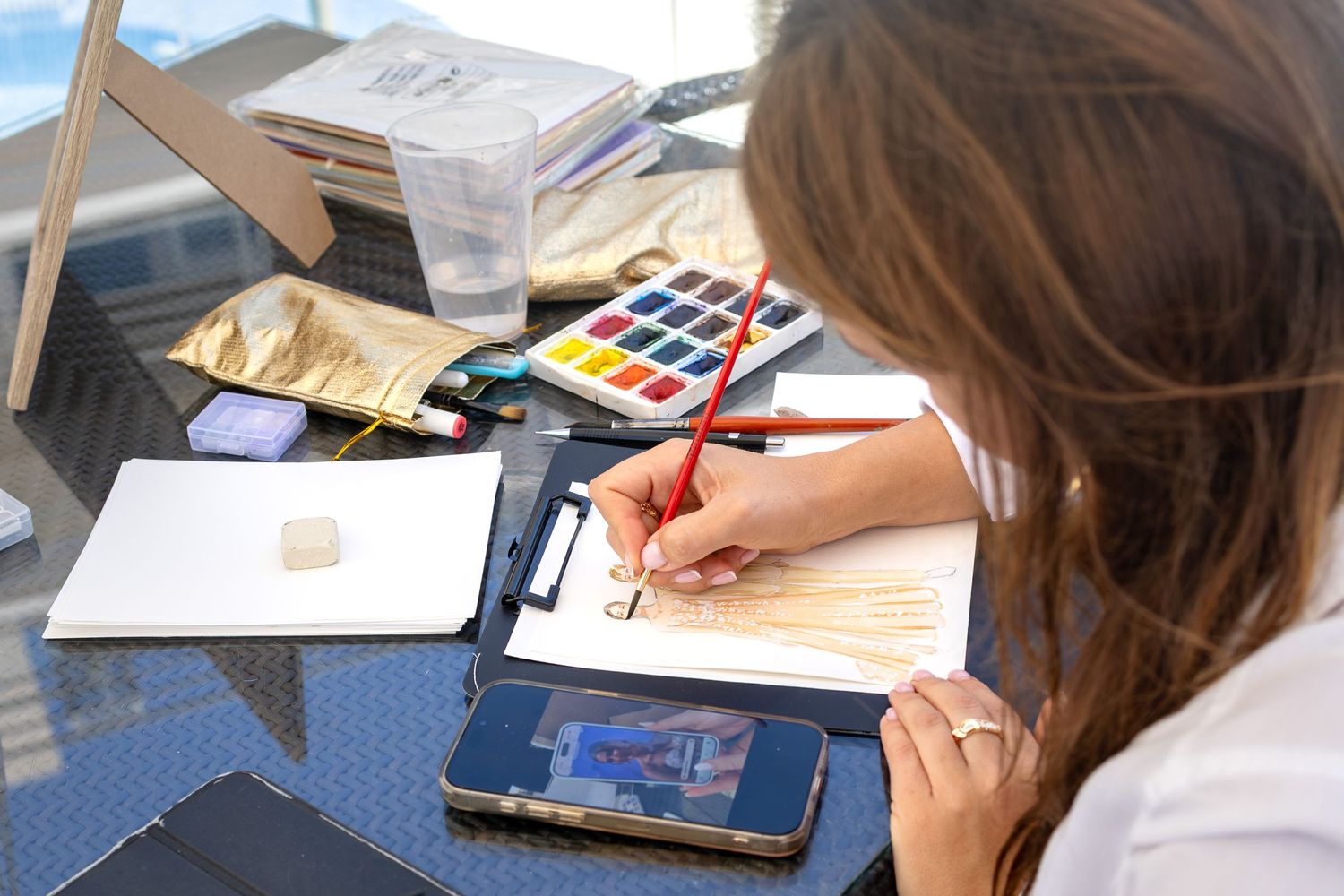 Woman painting wedding dress watercolor art at a desk with paints, brushes, and a tablet reference.