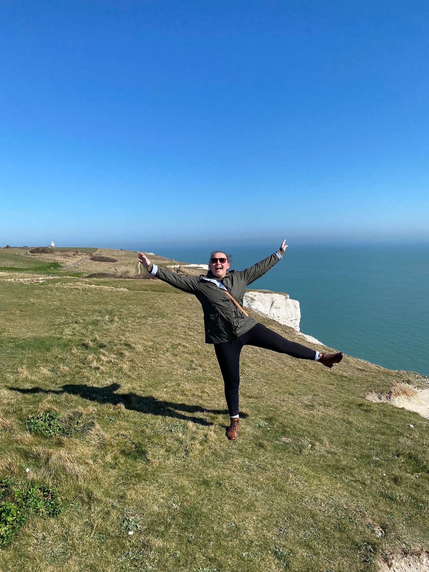 Person striking playful yoga poses on coastal clifftop against bright blue sky and ocean backdrop.