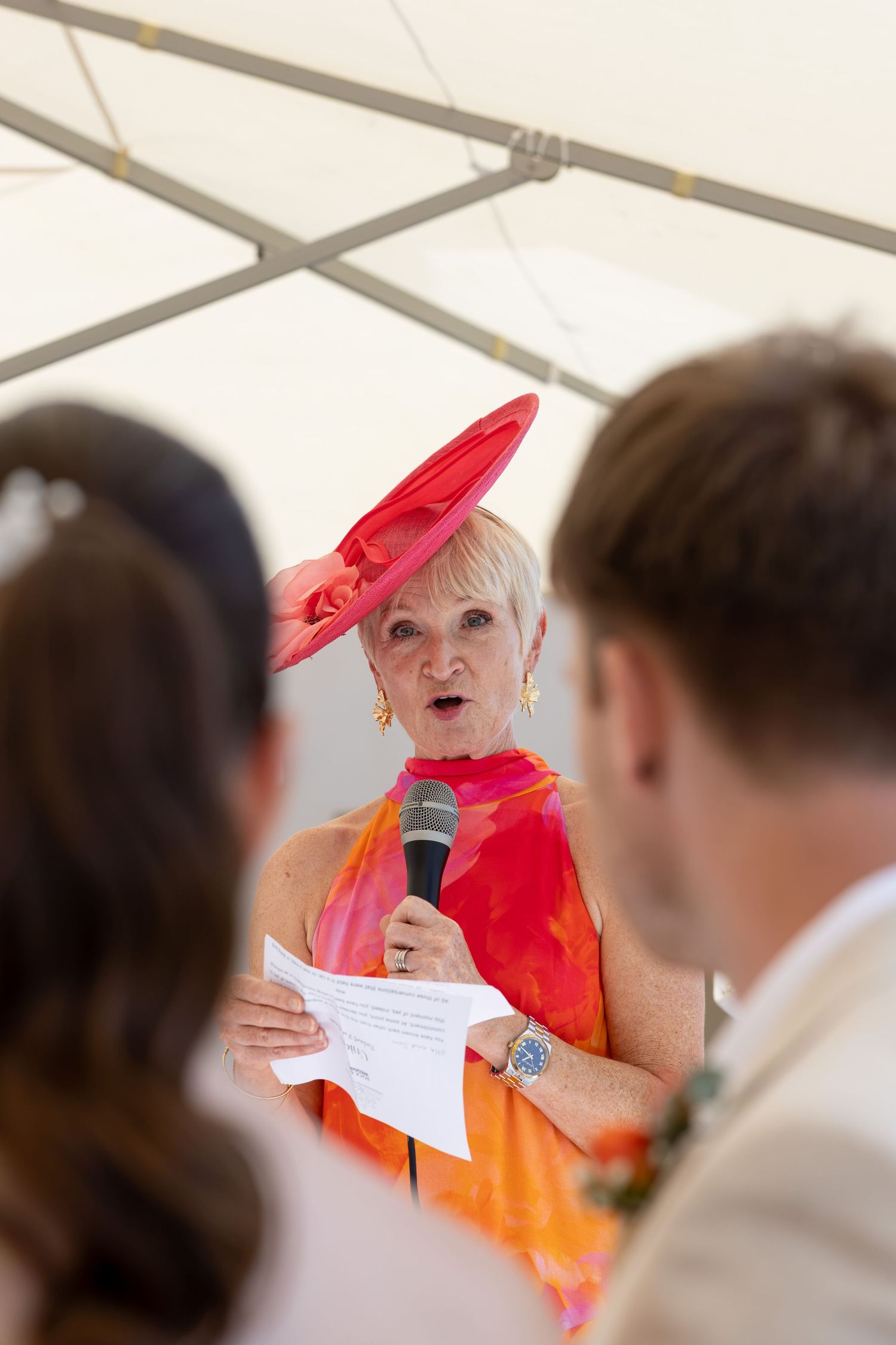 Elegant woman in red hat and colorful dress speaks into microphone at outdoor wedding ceremony.