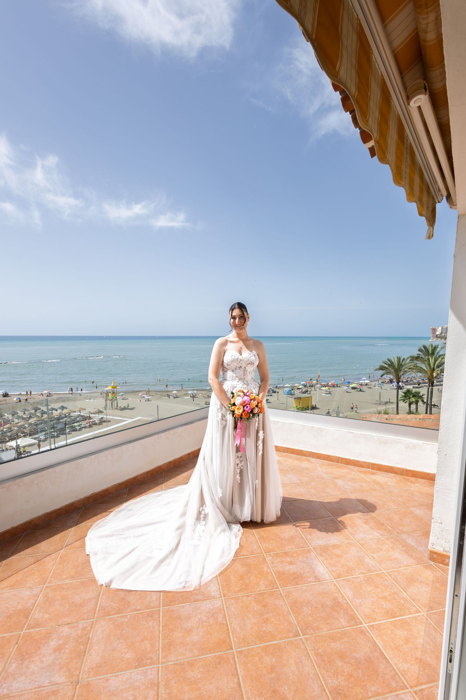 A bride in a flowing white lace gown holds a colorful bouquet on a sunny seaside balcony.