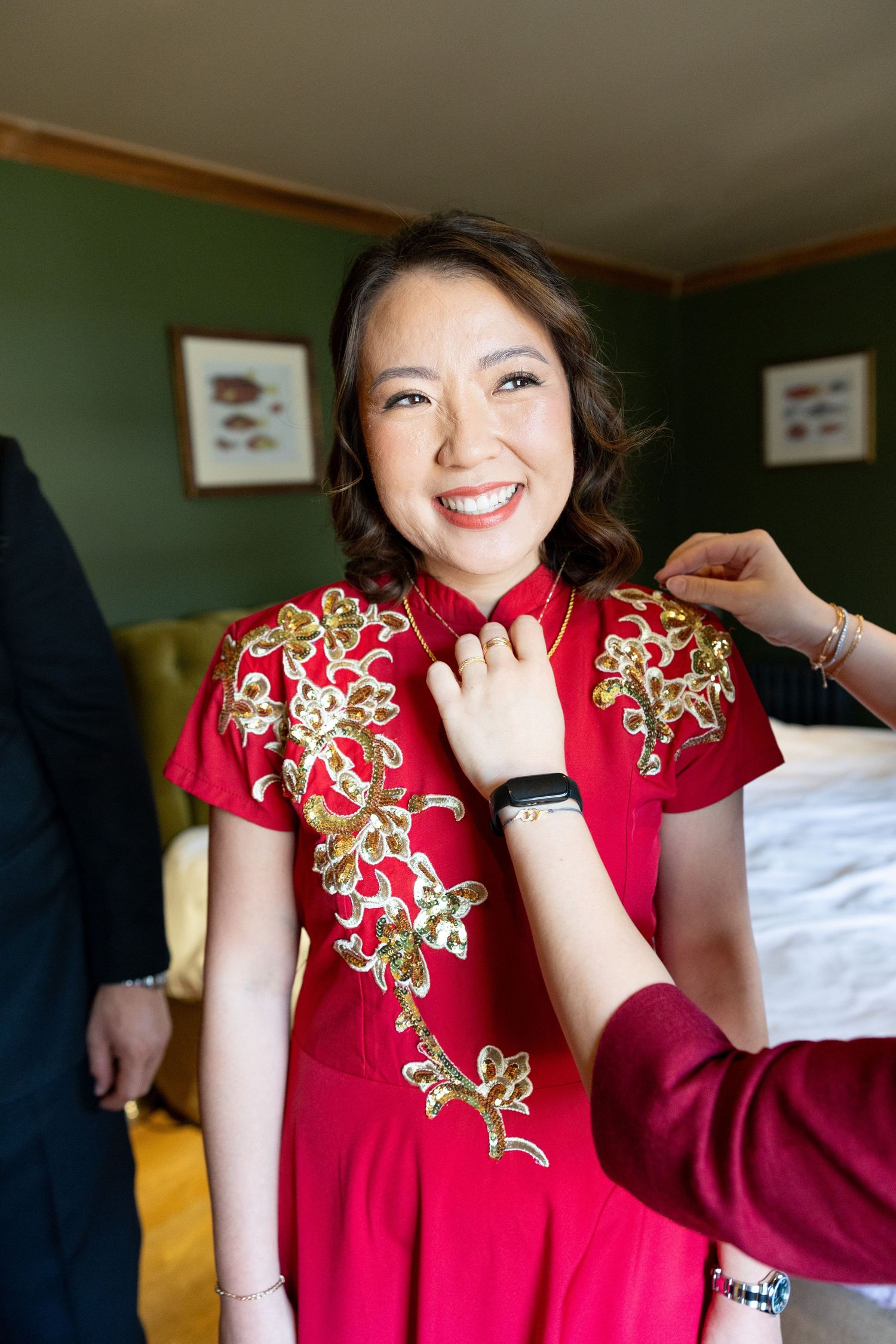 A smiling bride wearing a red and gold embroidered Chinese qipao dress being helped with her necklace.